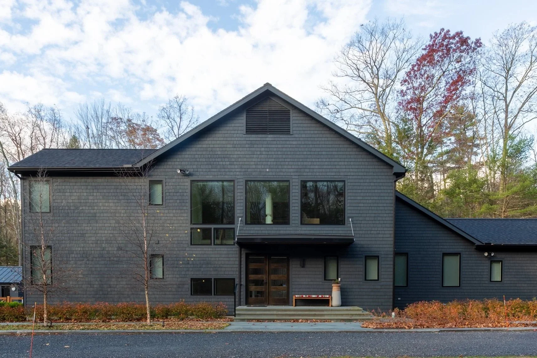 A modern two-story house with dark gray siding, large windows, and a small balcony above the front door, set against a backdrop of leafless trees and a partly cloudy sky.