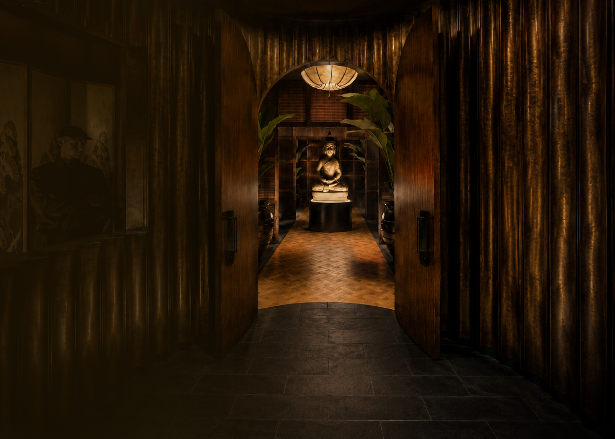Entrance to a dimly lit room with wooden walls and a statue of a seated person in the center, surrounded by large leafy plants, a hanging lamp above, and framed artwork on the sidewalls.
