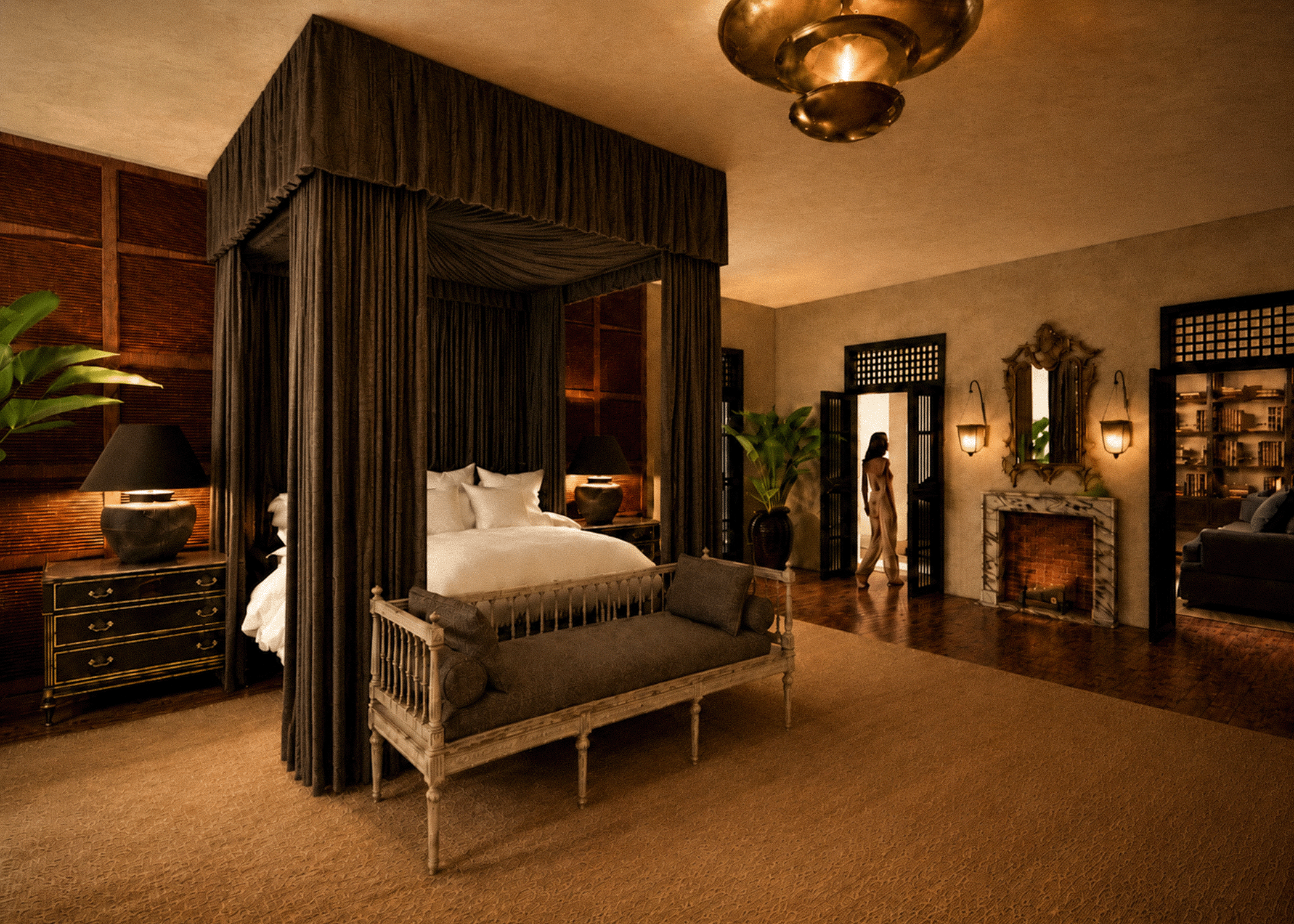 A luxurious hotel room with a canopy bed draped in dark curtains, beige walls, and warm lighting. Potted plants and vintage-style furniture, including a bench and nightstands, decorate the space. A woman stands near the doorway, and glowing lamps and a fireplace add to the cozy ambiance.