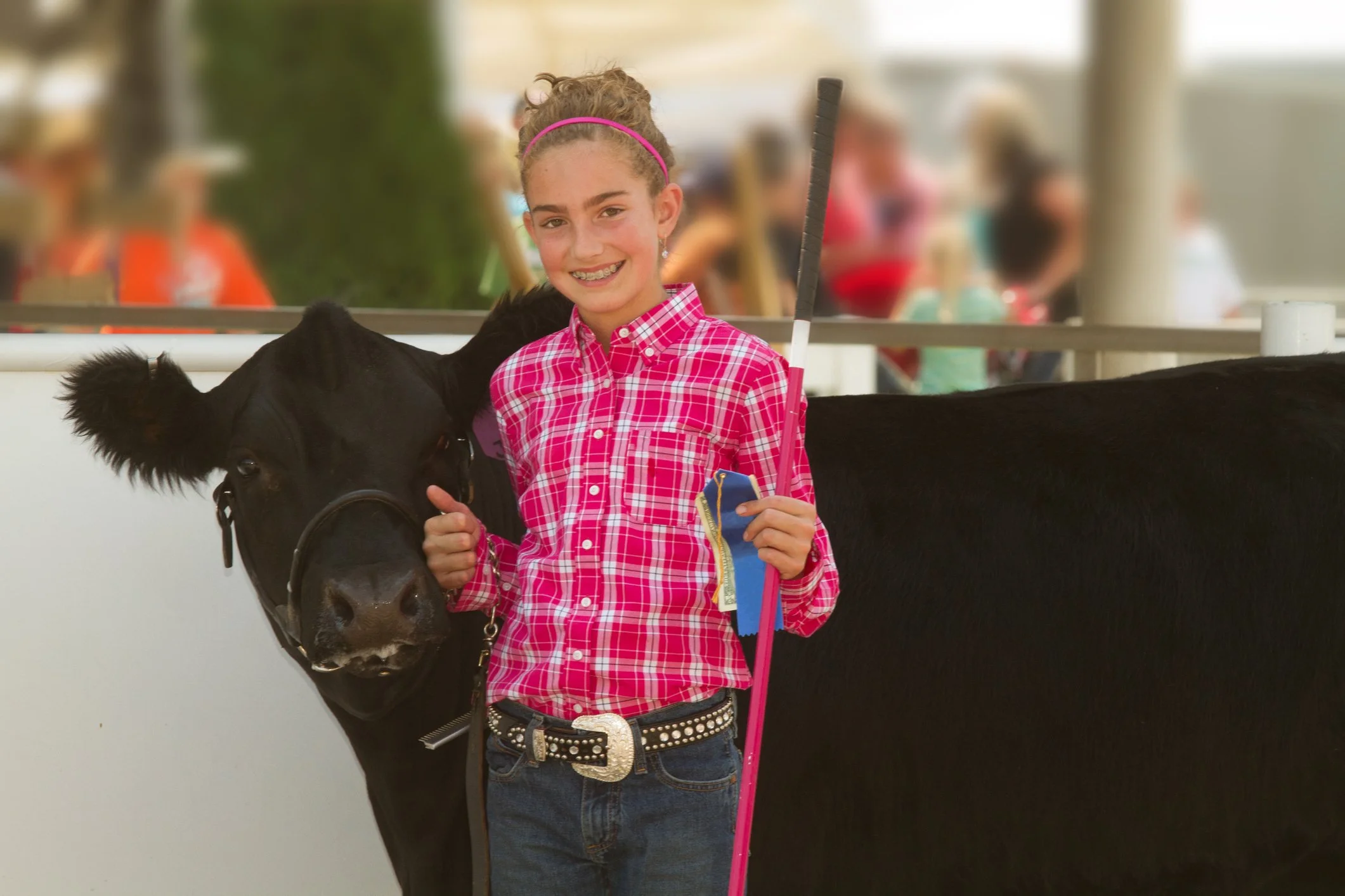 A young girl with braces wearing a pink checkered shirt, pink headband, and a decorative belt is standing next to a black horse, holding a pink stick and a blue ribbon. They are at an indoor or covered equestrian event with blurred spectators in the background.