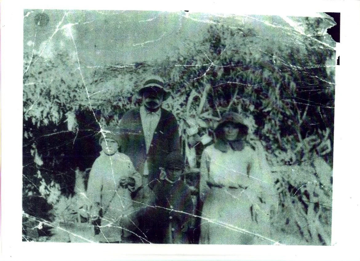 Vintage black and white photograph of a woman and three children outdoors near foliage, with visible creases and damage to the photo.