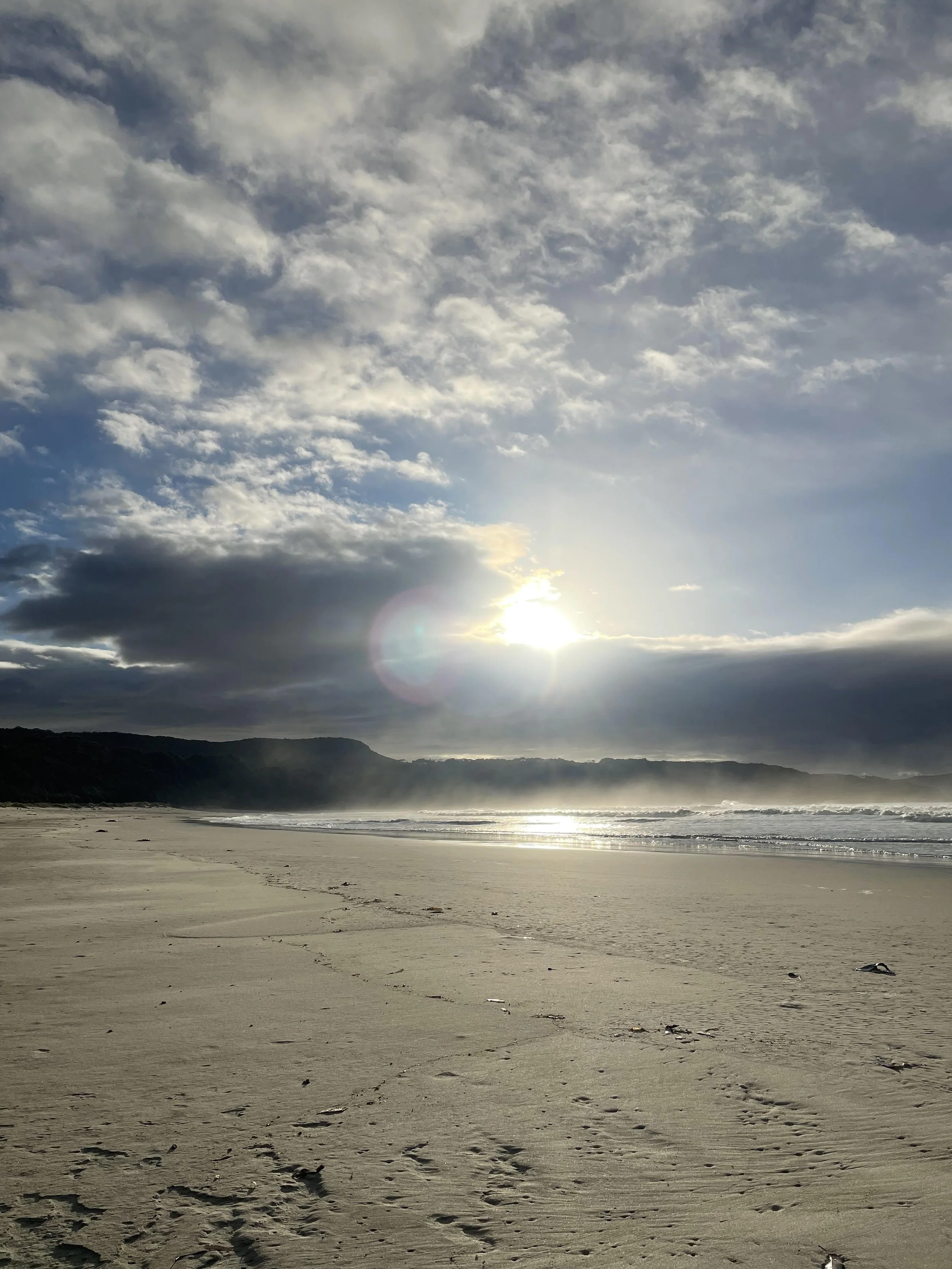 Sunset over a sandy beach with footprints, cloudy sky, and distant cliffs.