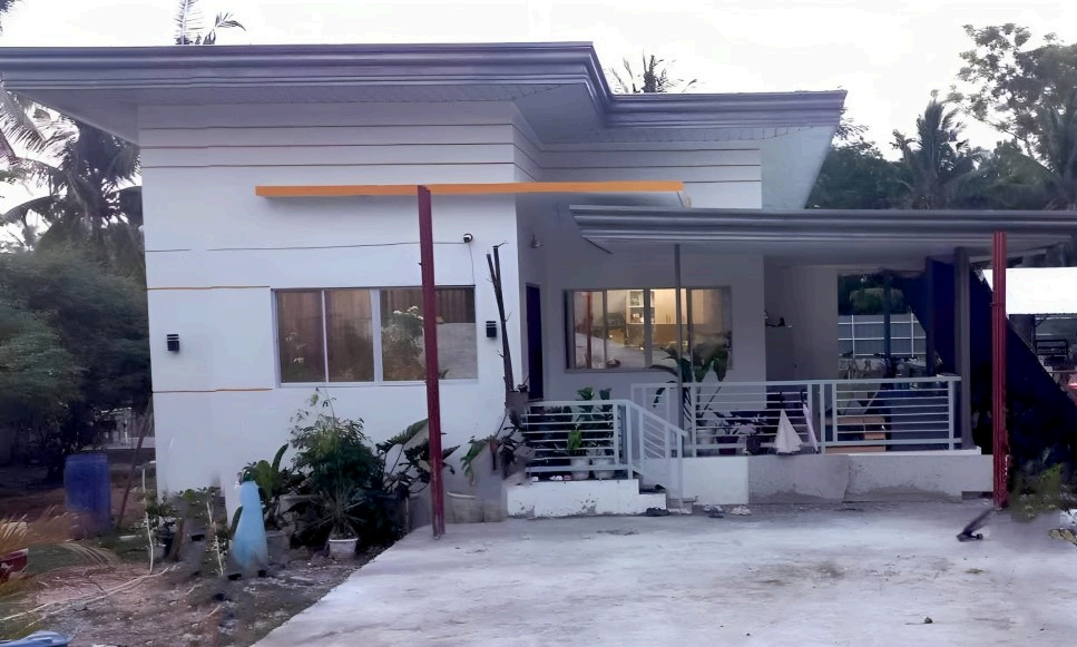 Modern white house with a small front yard, plants, and a separated porch area with a railing, some chairs, and umbrellas, set amid greenery with trees in the background.
