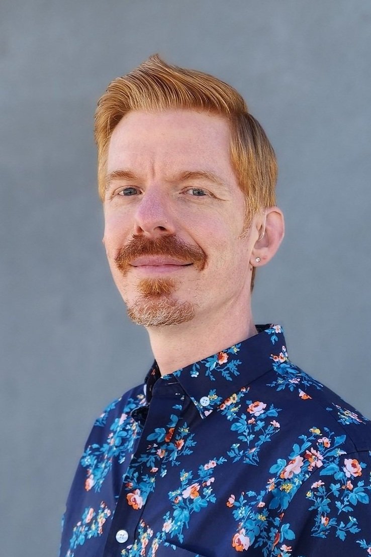  Picture of Greg Anderson, a man with red hair and a goatee, in a floral shirt, smiling and looking directly at the viewer. 