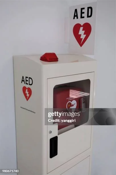 AED emergency defibrillator cabinet with a red AED device inside, marked with a heart and lightning bolt symbol, located in a white cabinet on a white wall.