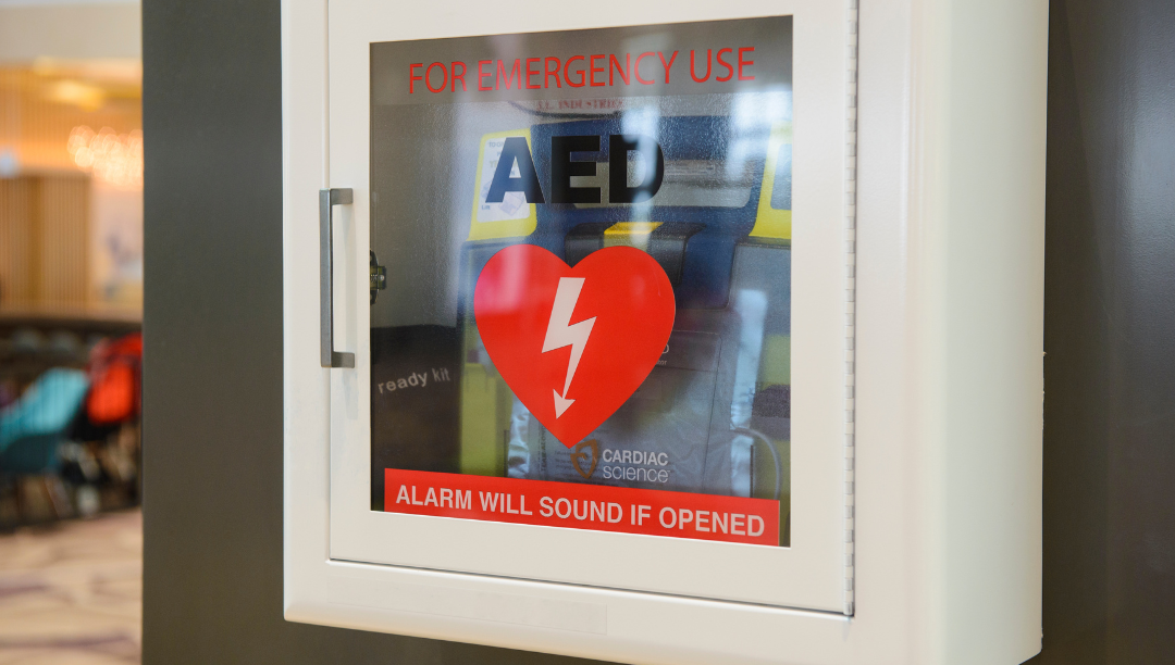 White emergency AED cabinet with a glass door, red warning labels, and a heart symbol with a lightning bolt, indicating it is for emergency use and the alarm will sound if opened.