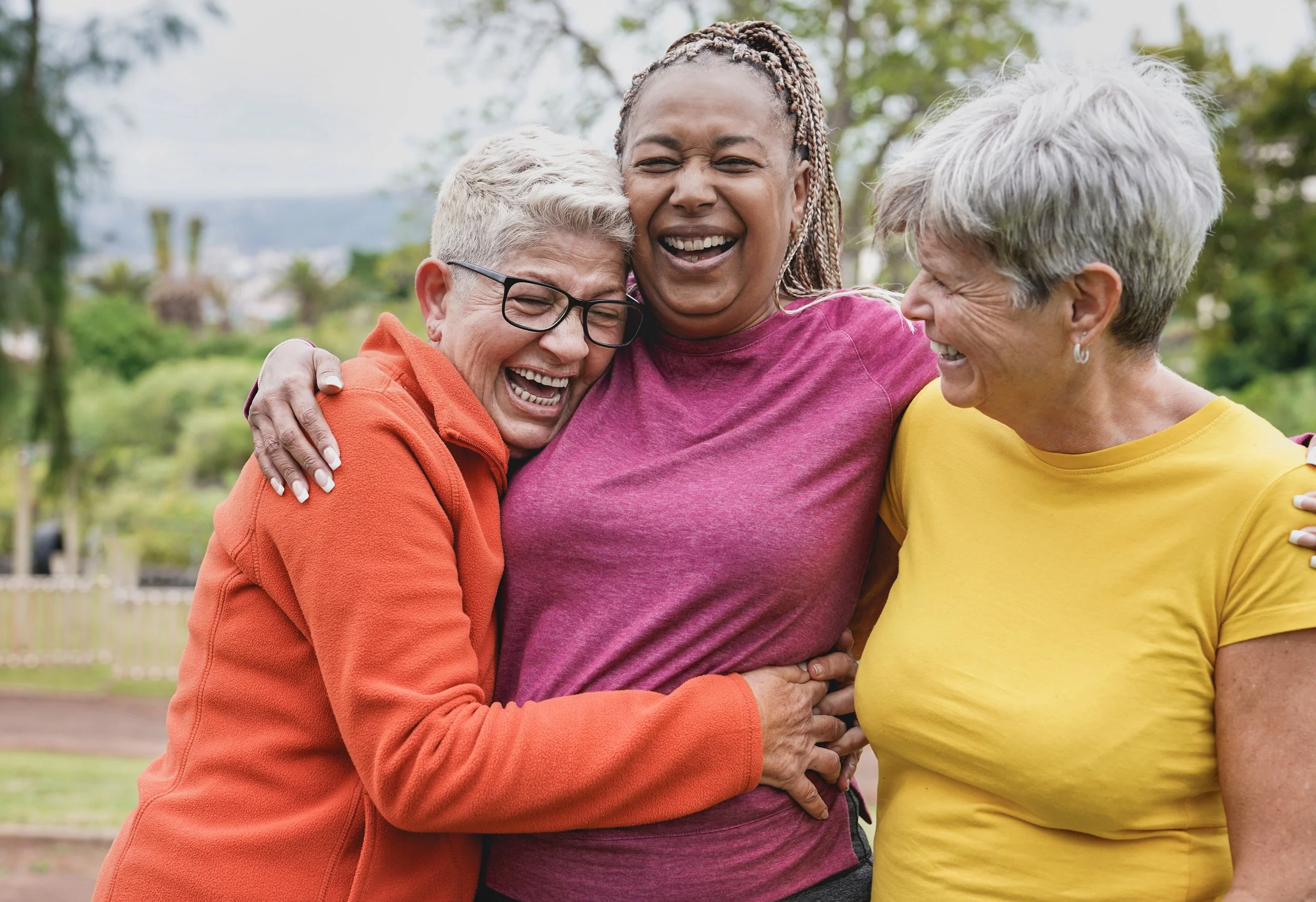 Three older women laughing and hugging outdoors, wearing colorful casual clothing.