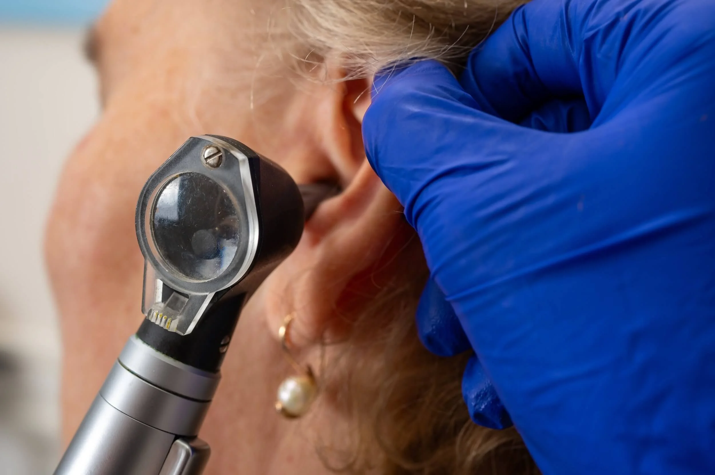 A healthcare professional wearing a blue glove examining an elderly patient's ear using an otoscope.