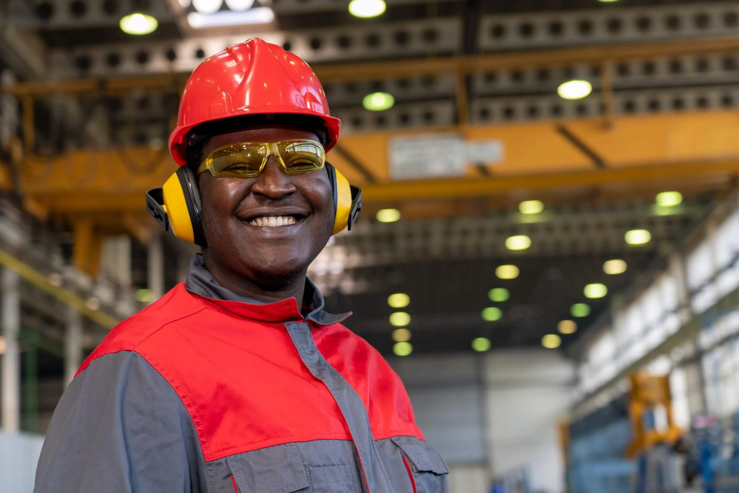 A smiling man in a red and gray safety uniform, wearing a red safety helmet, yellow ear protection, and safety glasses, standing in an industrial warehouse or factory setting.