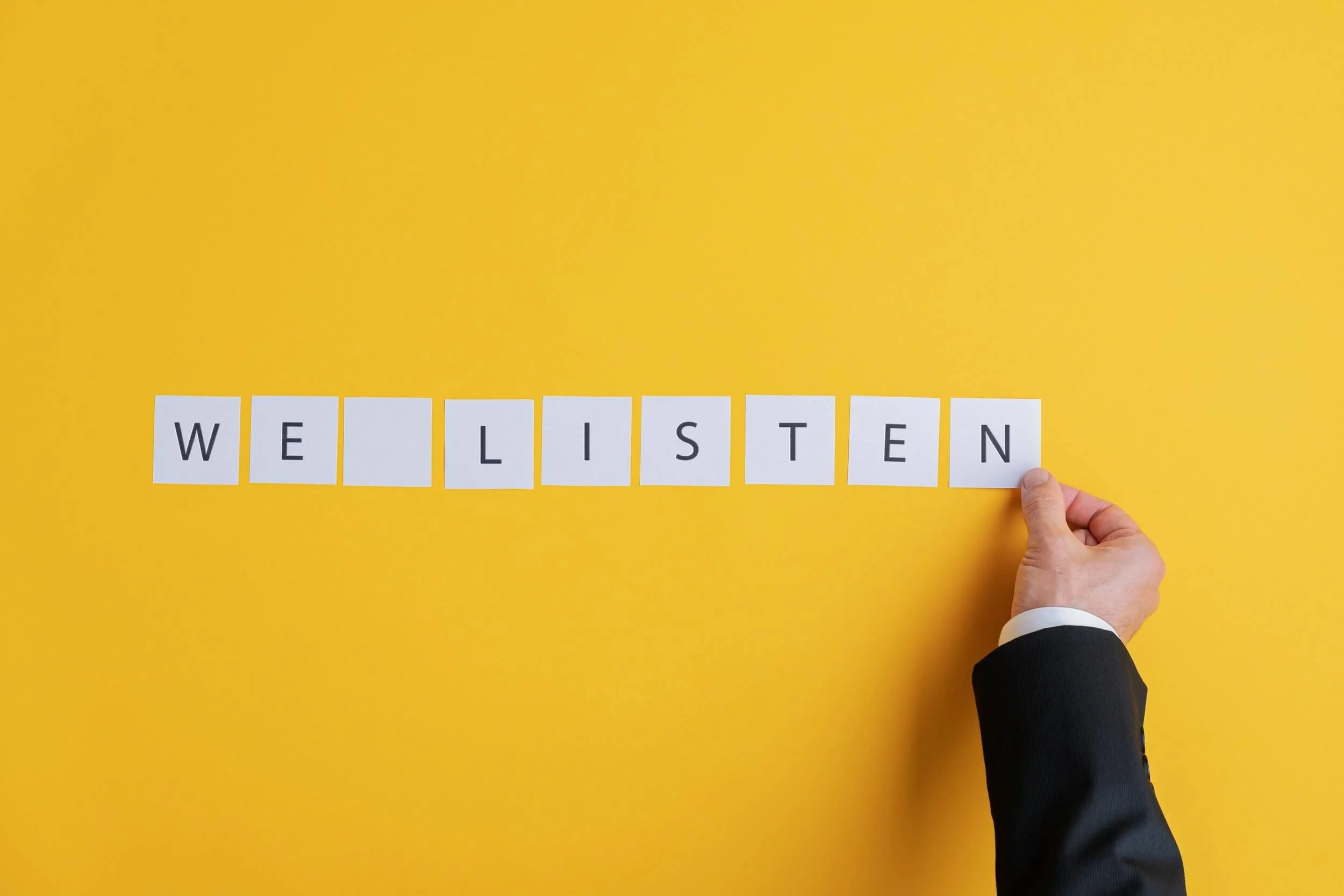 Hand holding a letter 'N' tile to complete the words 'WE LISTEN' on a yellow background.