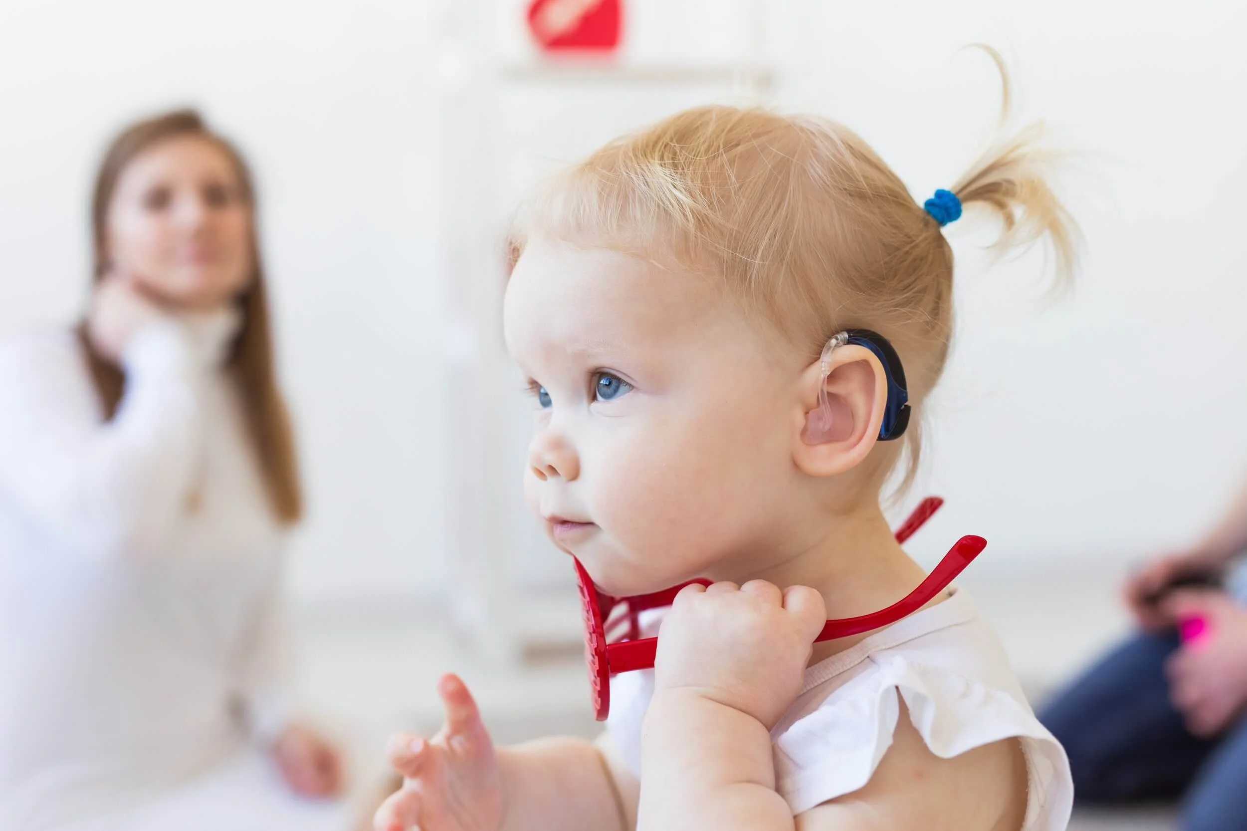 A young girl with blonde hair and blue eyes, wearing a hearing aid, sitting at a medical or clinical setting. She is holding a red stethoscope around her neck. In the background, a woman, possibly a healthcare professional, is blurred.