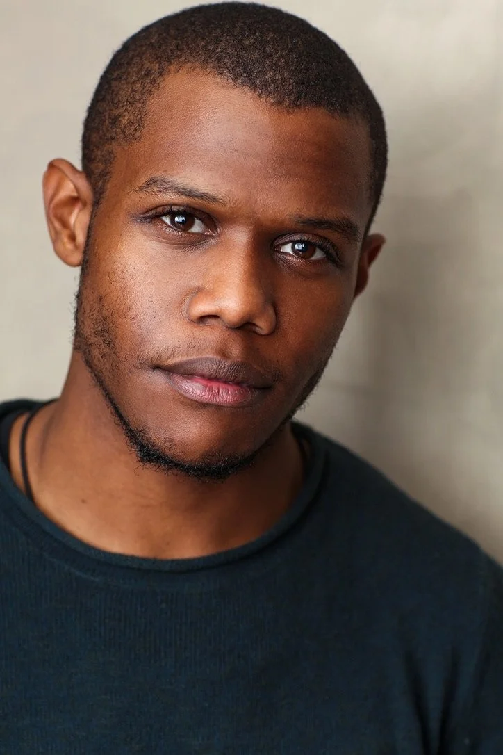 Close-up portrait of a young man with short hair and a slight beard, wearing a black shirt, looking directly at the camera with a neutral expression.