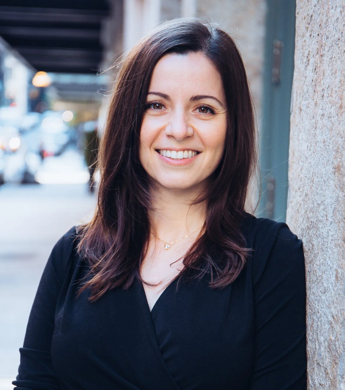 A woman with dark brown hair smiling, standing outdoors against a stone wall with blurred cars and buildings in the background.