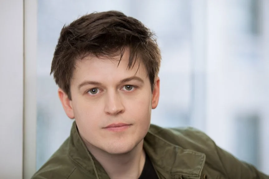 Close-up of a young man with short brown hair, fair skin, and wearing a green jacket, standing indoors in front of a blurred background.
