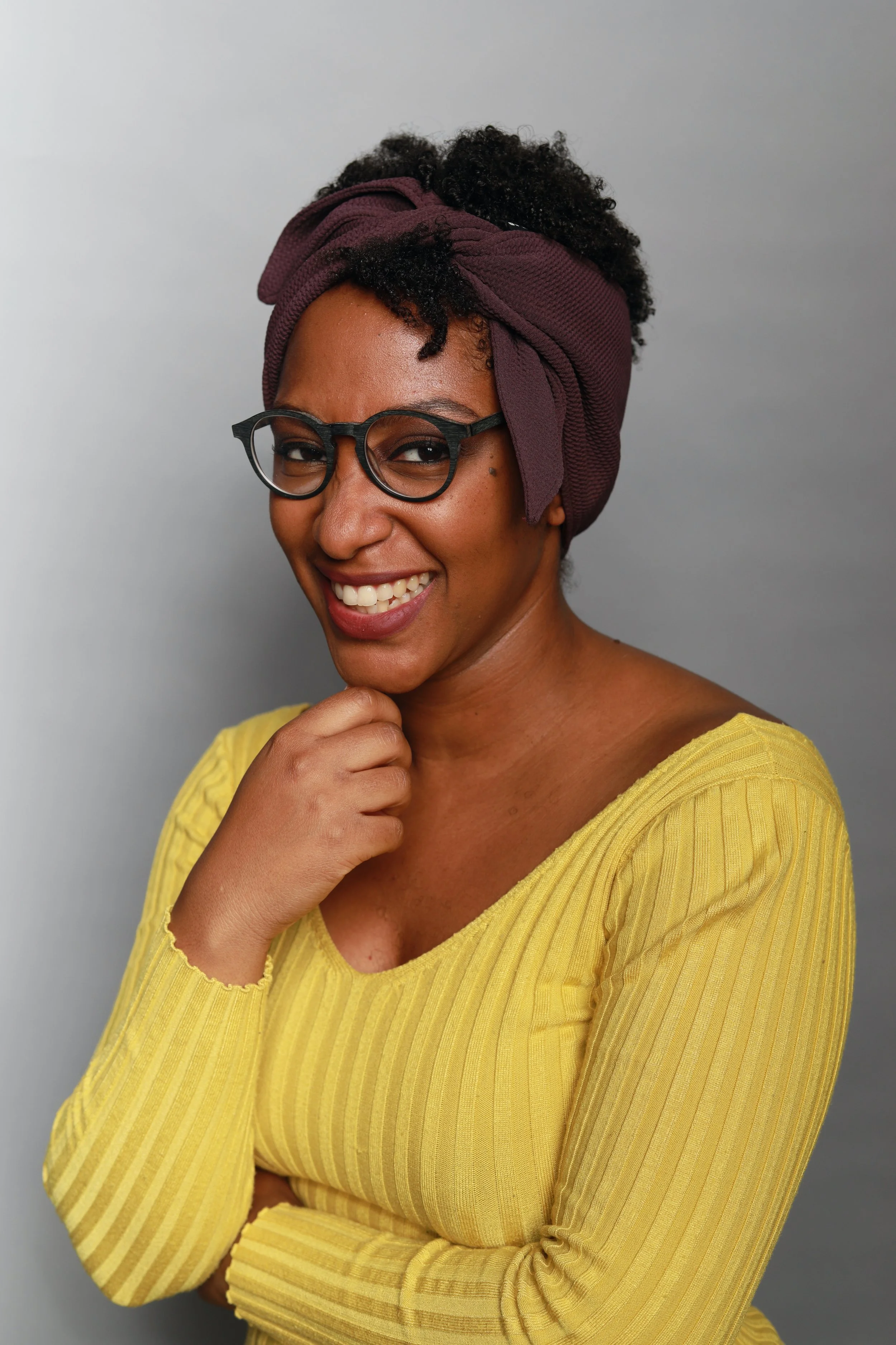 A smiling woman wearing glasses, a purple headwrap, and a yellow ribbed top, standing against a gray background.