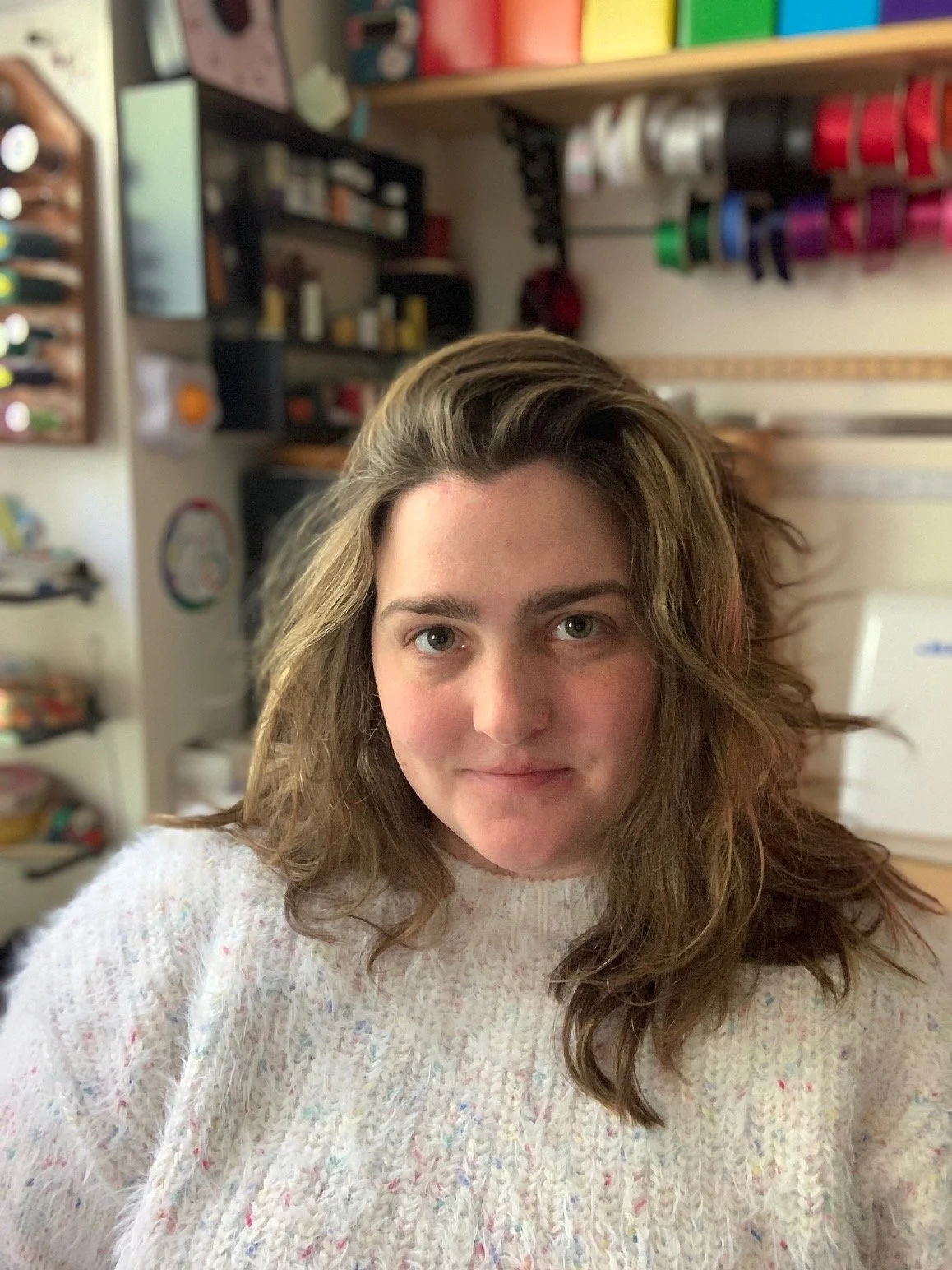 A woman with wavy hair wearing a light-colored sweater, indoors with shelves of craft supplies in the background.