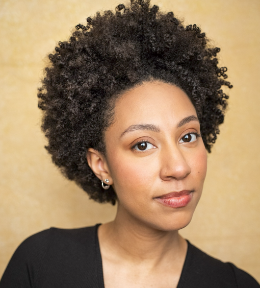 A young woman with curly black hair and light brown skin, wearing small earrings and a black top, looking at the camera with a neutral expression against a beige background.