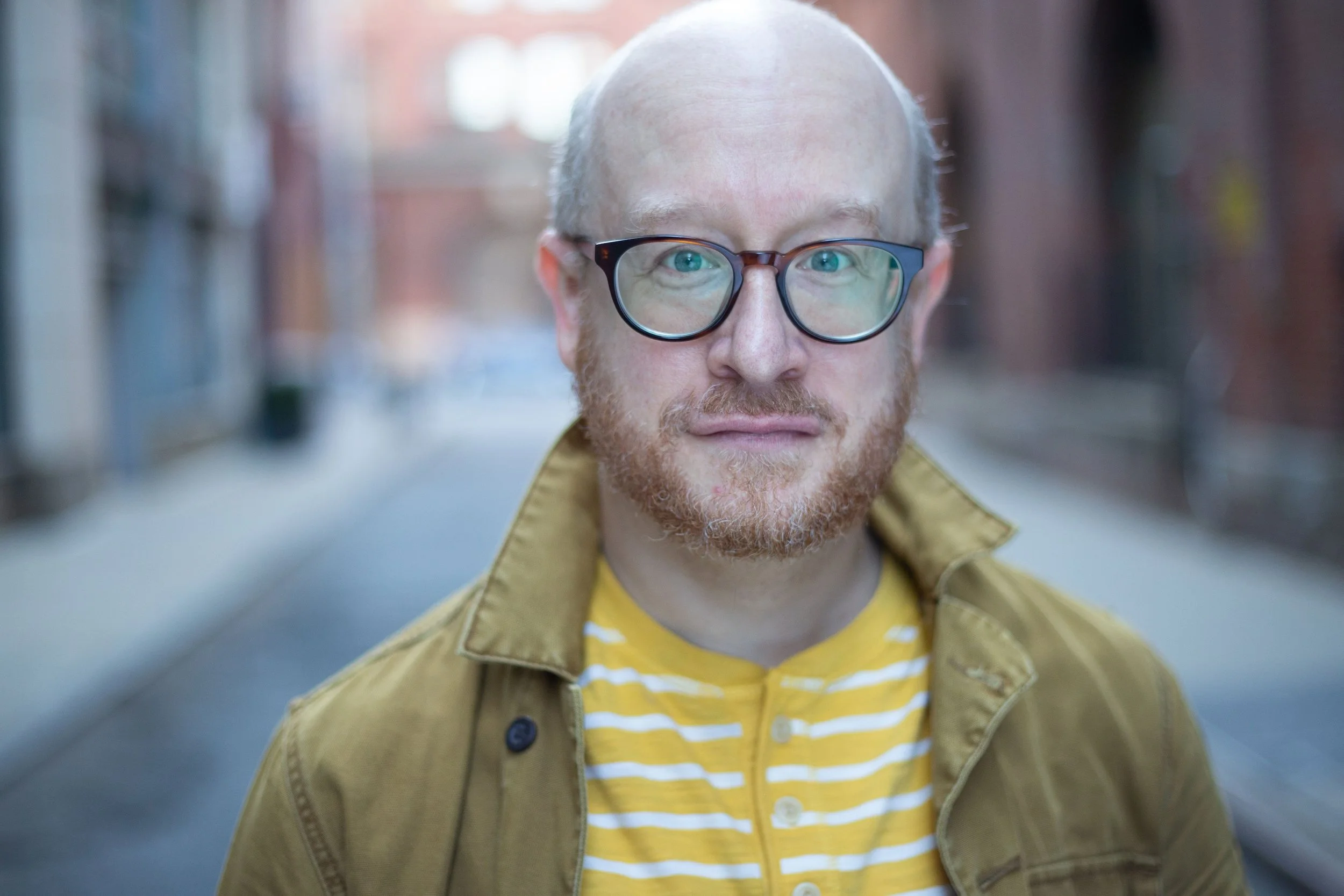 A man with glasses, a beard, and a shaved head standing on a city street during daytime, wearing a yellow and white striped shirt and a brown jacket.