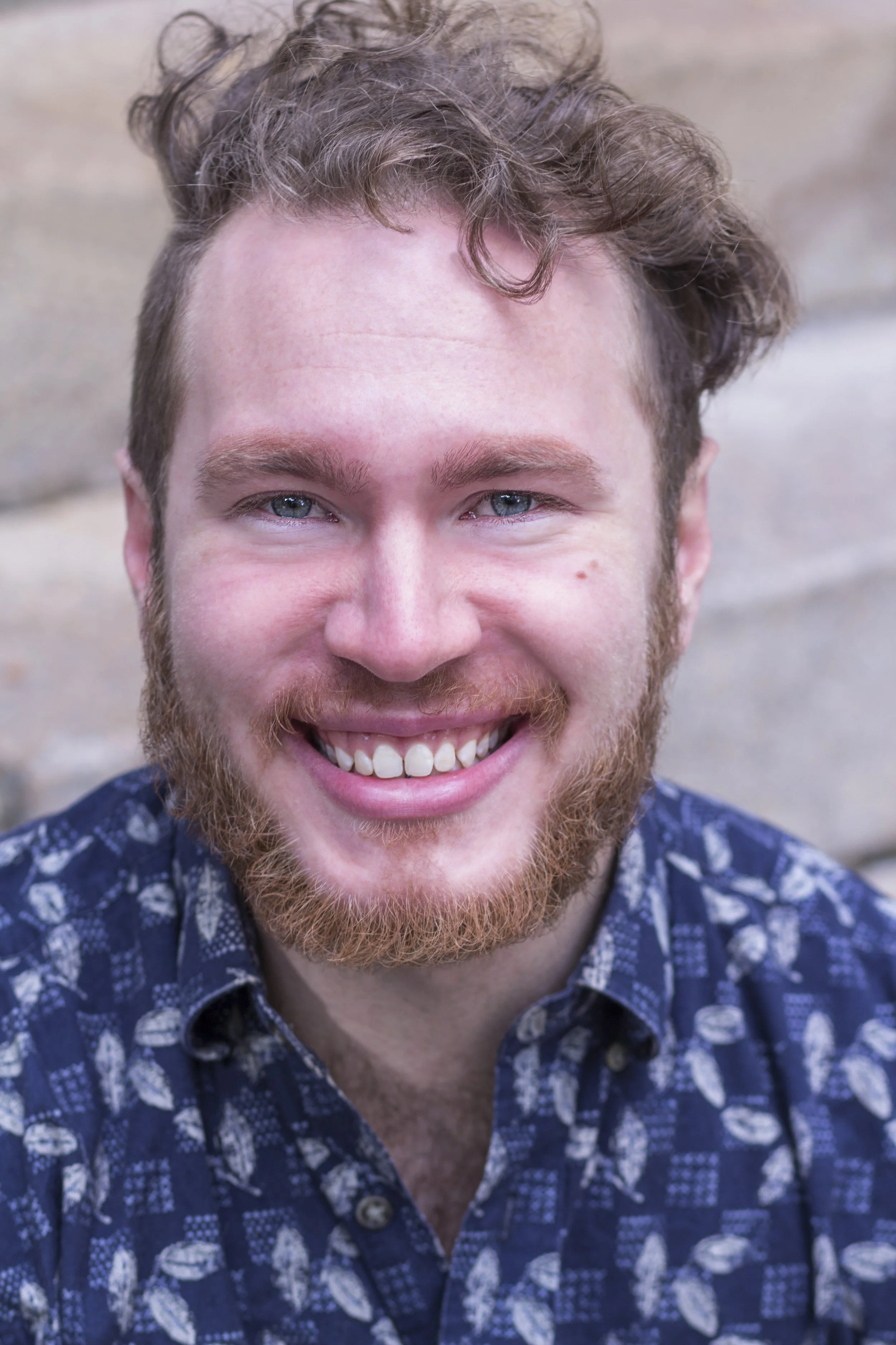 Close-up portrait of a smiling young man with curly brown hair, blue eyes, and a reddish beard, wearing a patterned shirt, standing outdoors against a stone wall.