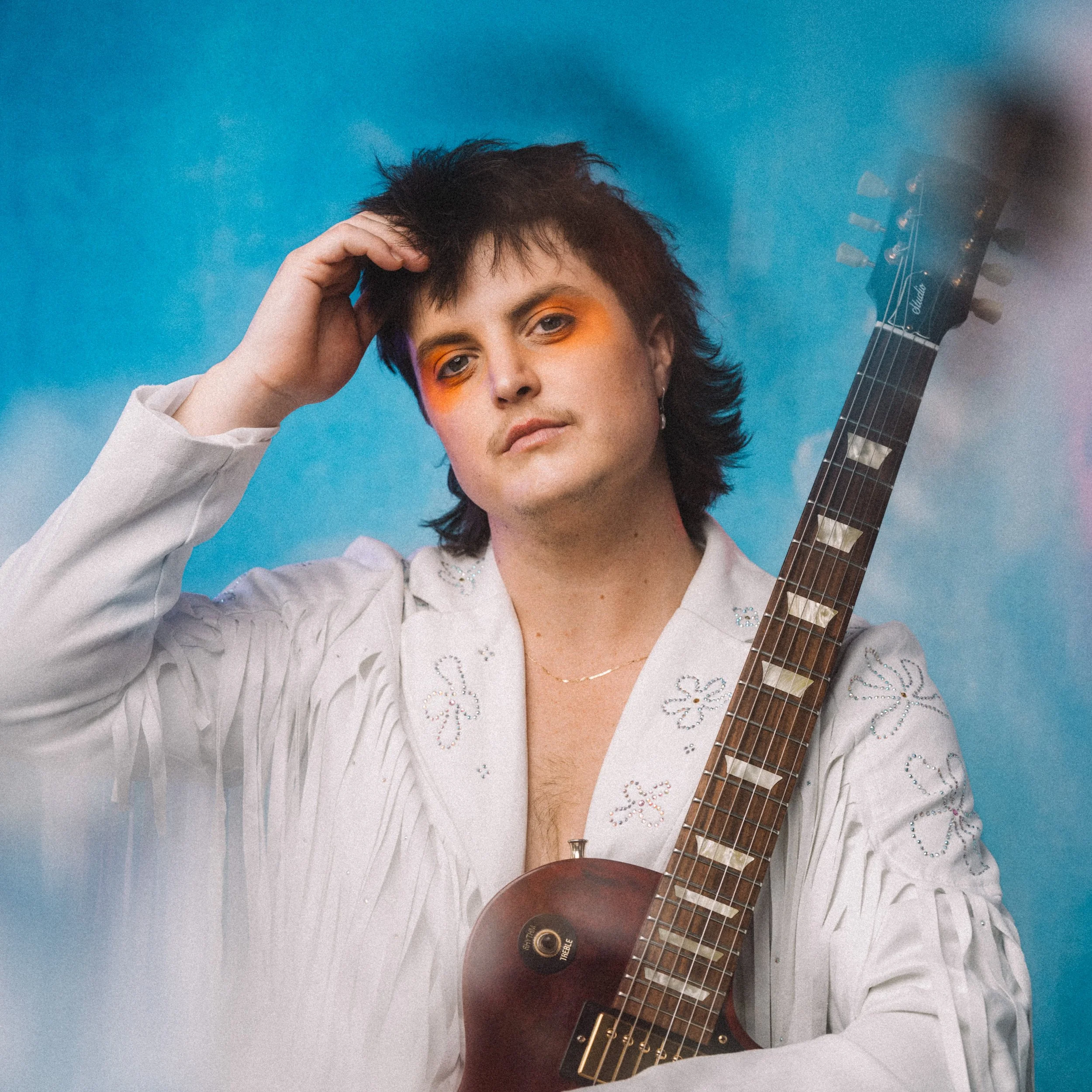 A person with dark, short hair and orange eye makeup holding a guitar, wearing a white embroidered jacket, against a blue background.