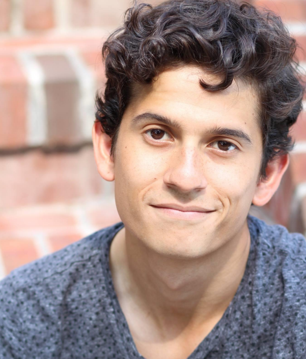 Close-up of a young man with curly brown hair smiling softly, wearing a gray textured shirt, against a blurred brick wall background.