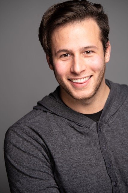 A young man with brown hair and a beard smiling, wearing a gray hoodie, against a plain gray background.