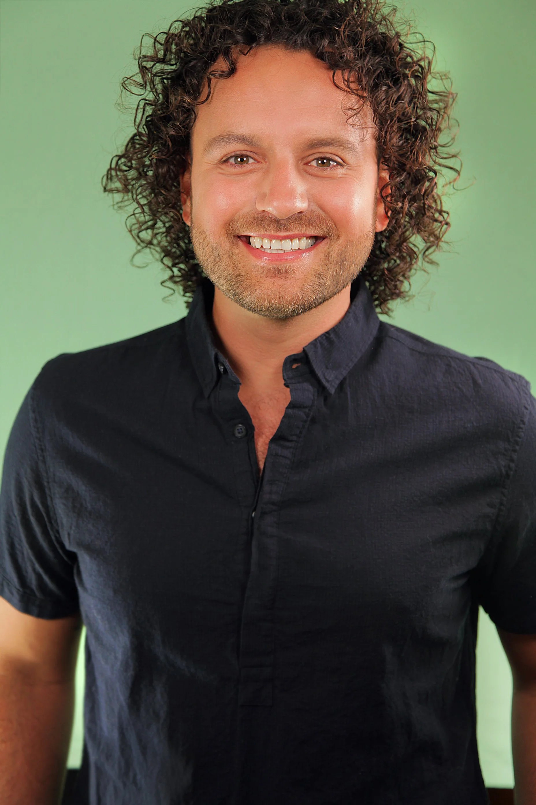 Portrait of a smiling man with curly hair, wearing a black shirt, standing in front of a green background.