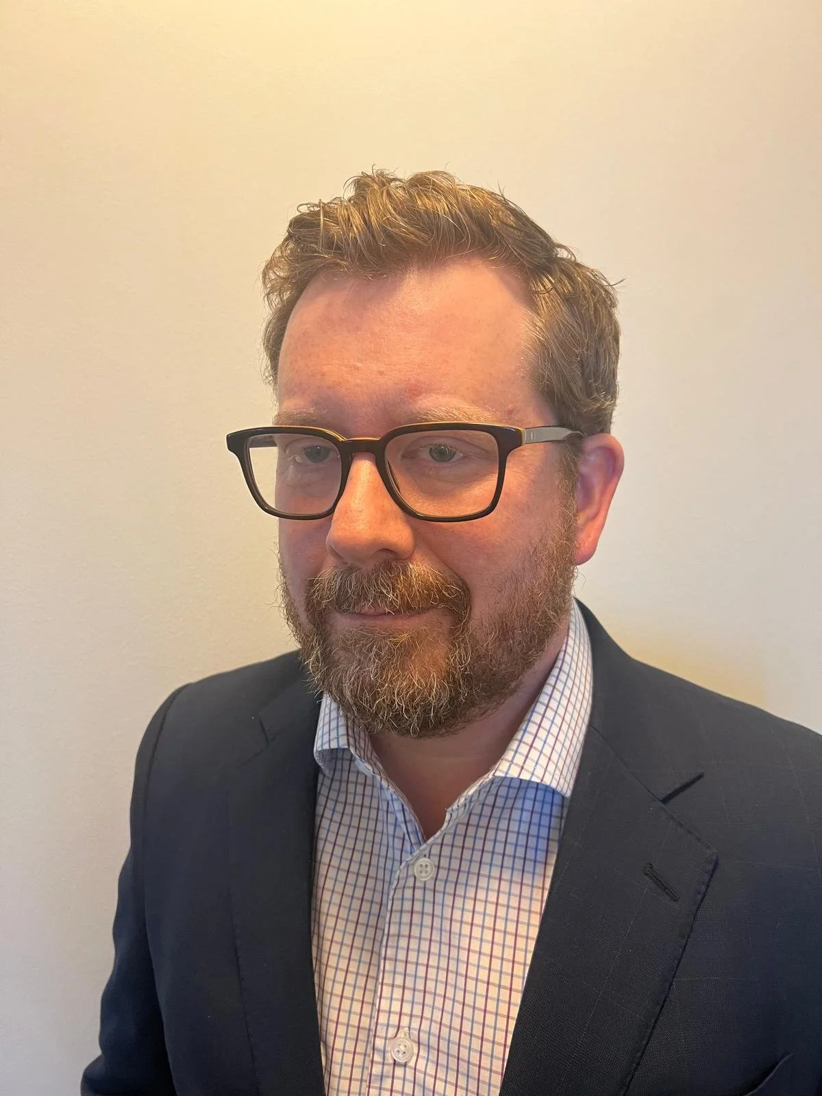 A man with glasses, a beard, and short curly hair wearing a dark suit and a checkered shirt stands against a plain white wall.