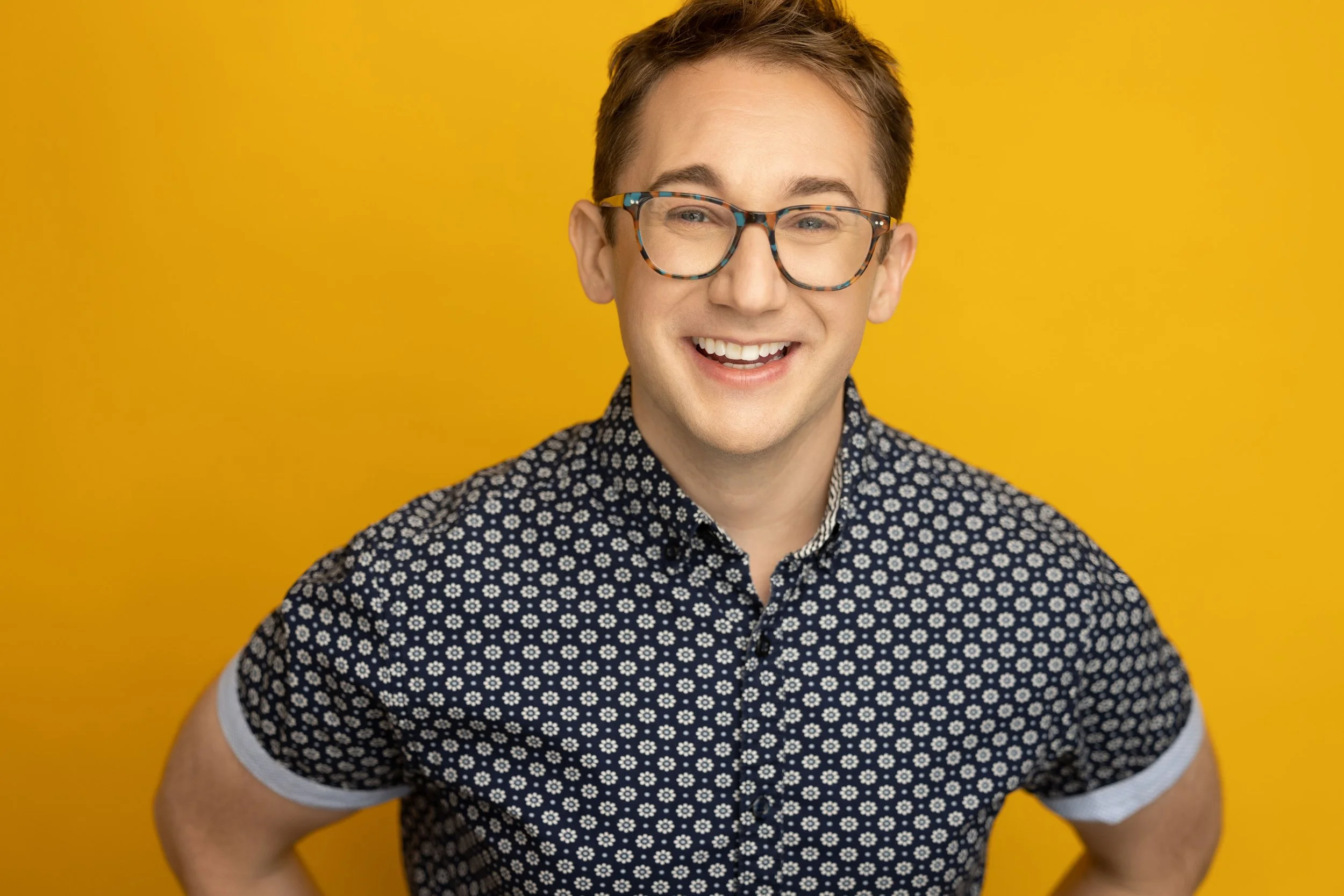 Young man with glasses smiling against a yellow background.