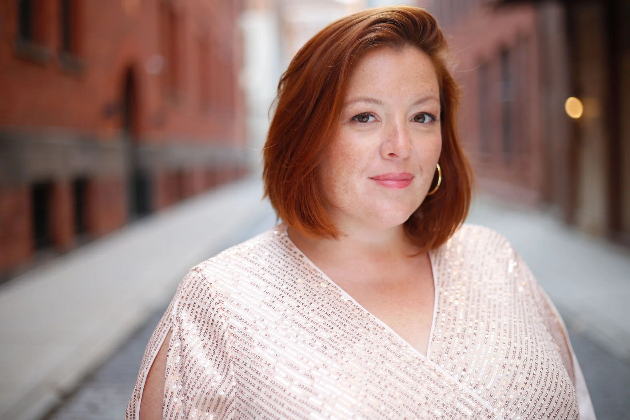 A woman with shoulder-length red hair, wearing a light pink, glittery top, stands in an alleyway with red brick buildings on either side.