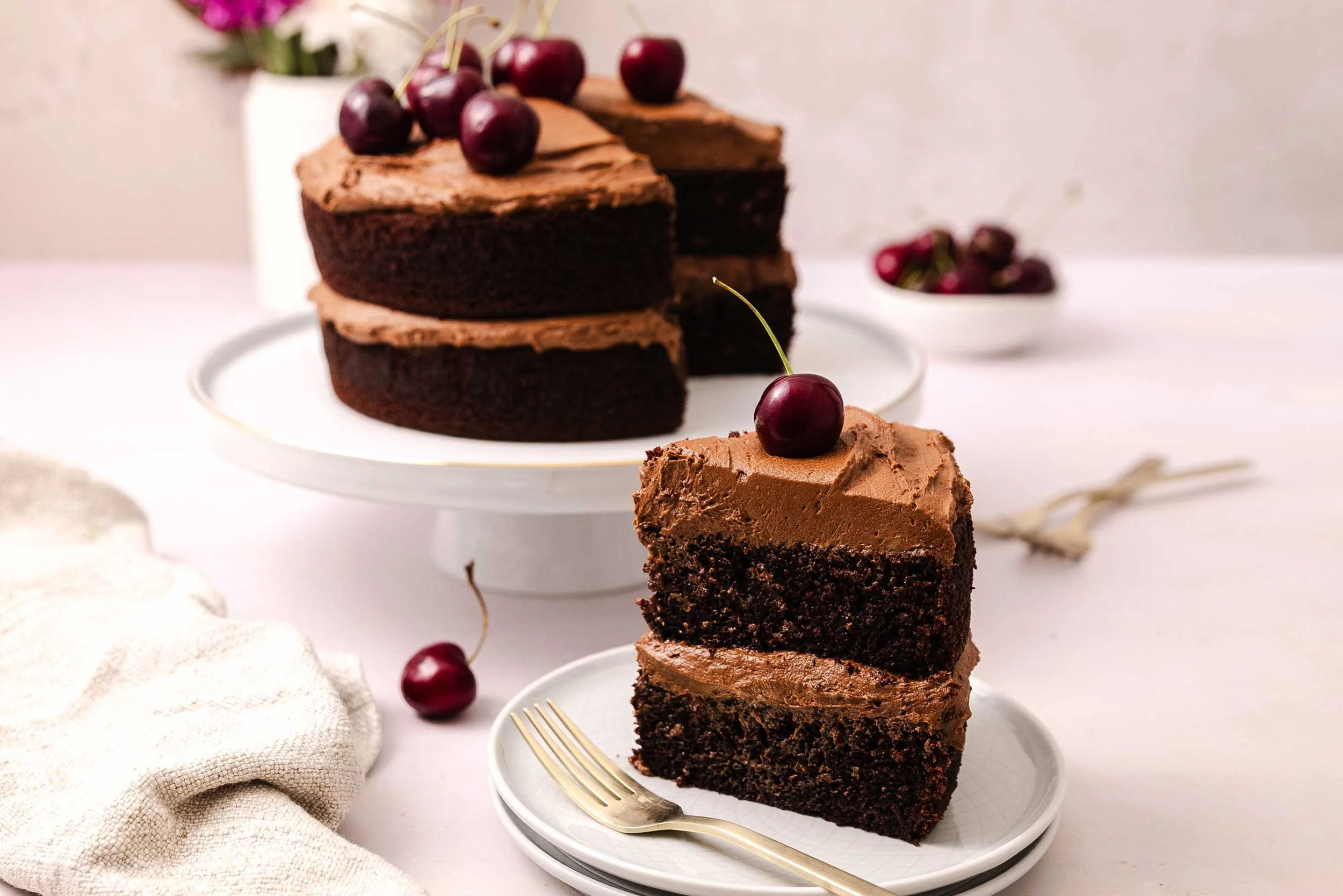 A slice of Double Layer Chocolate Cake, topped with a cherry and serves on a white plate