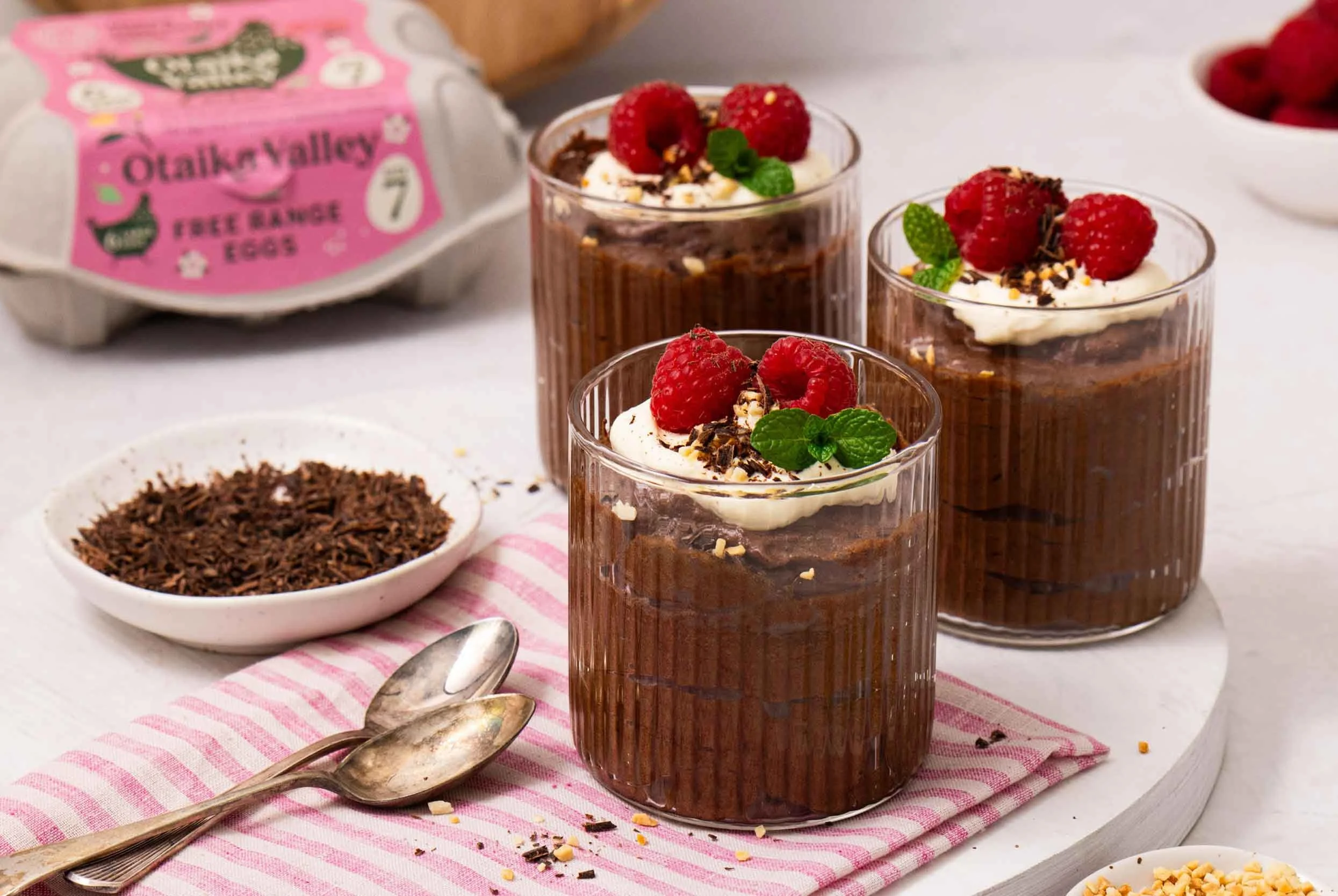 Three glass cups filled with chocolate mousse, and topped with cream, mint leaves, raspberries and shaved dark chocolate, served on a white wooden board, with two silver spoons and a Otaika Valley Free Range Egg carton in the background