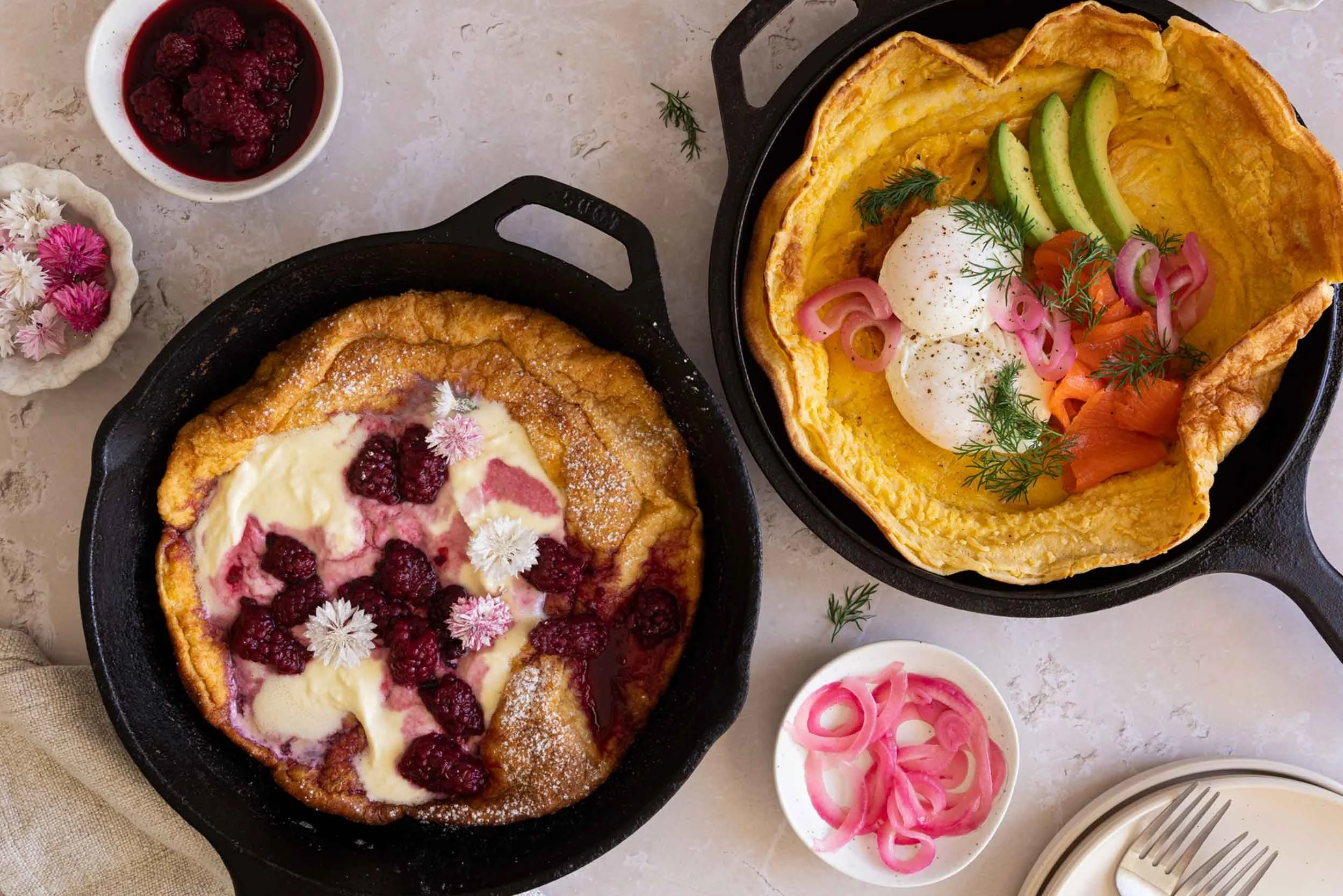 Two cast iron frying pans filled with Baby Dutch Pancakes, one topped with boysenberries and cream and the other topped with smoked salmon, avocado and poached egg