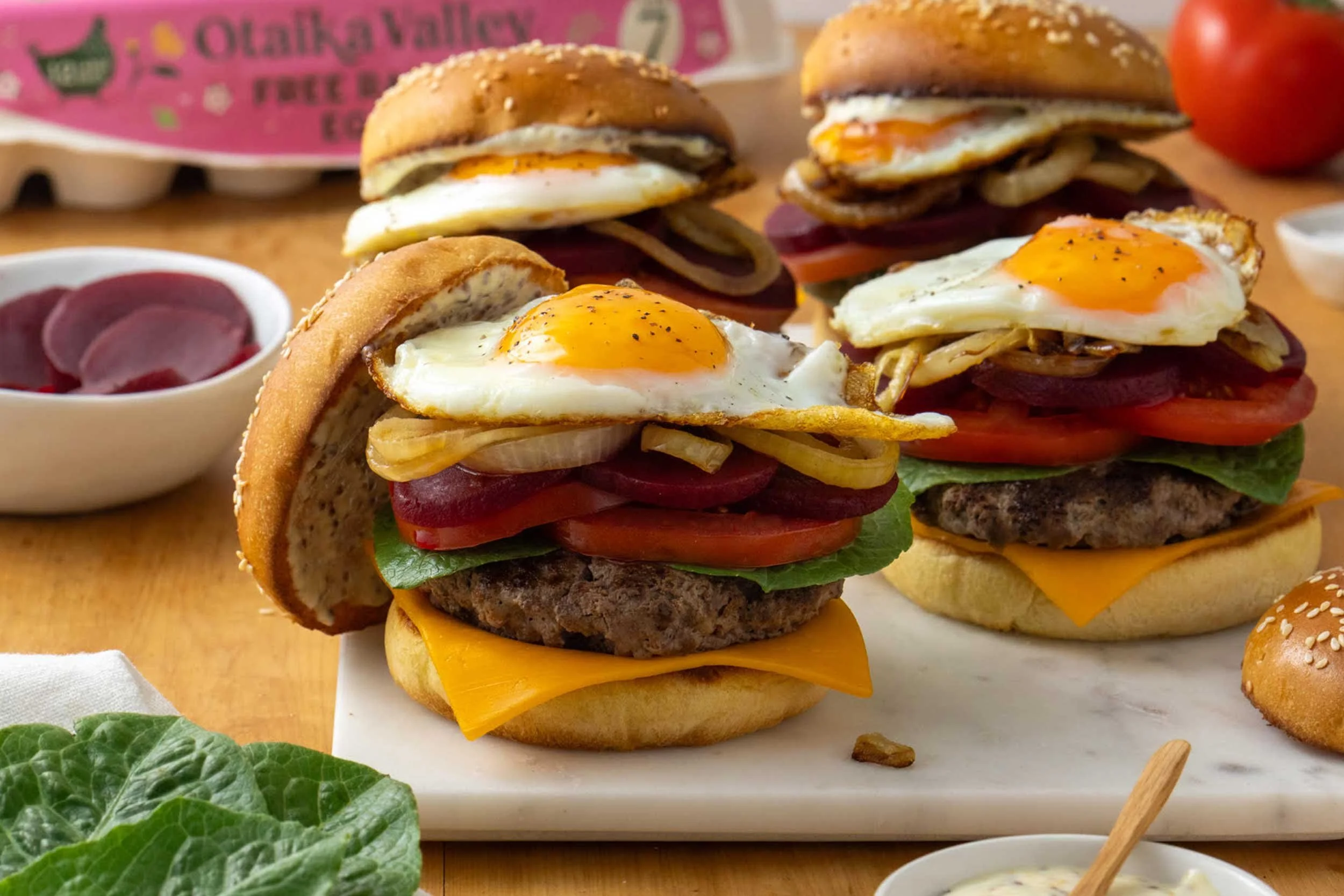 Four beef burgers, served on a marble tray, each featuring a beef patty, cheese, lettuce, tomato, beetroot, caramelised onion and a fried Otaika Valley Free Range egg