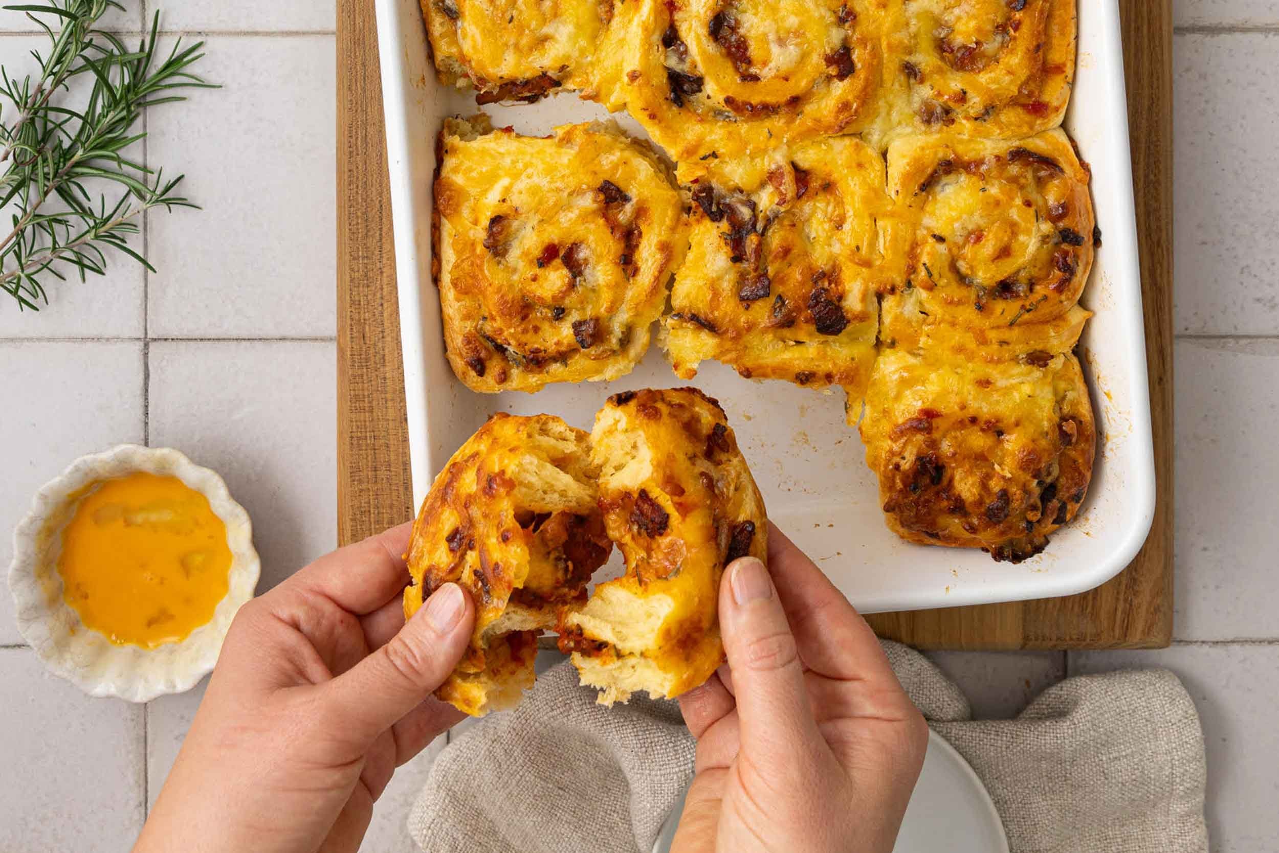 A baking dish filled with cheesy bacon and rosemary brioche scrolls, lightly golden. Two scrolls are missing, at the bottom of the image is two hands pulling apart one scroll.