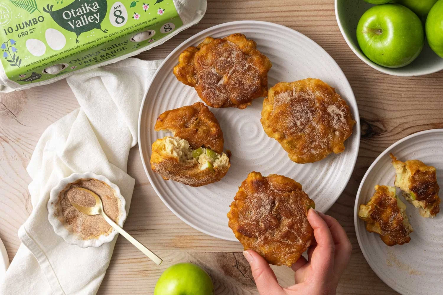 A white, round serving plate with four apple fritters on it, an Otaika Valley Free Range Egg carton in the top left corner, a small bowl filled with cinnamon sugar in the bottom left corner and Granny Smith apples in a bowl