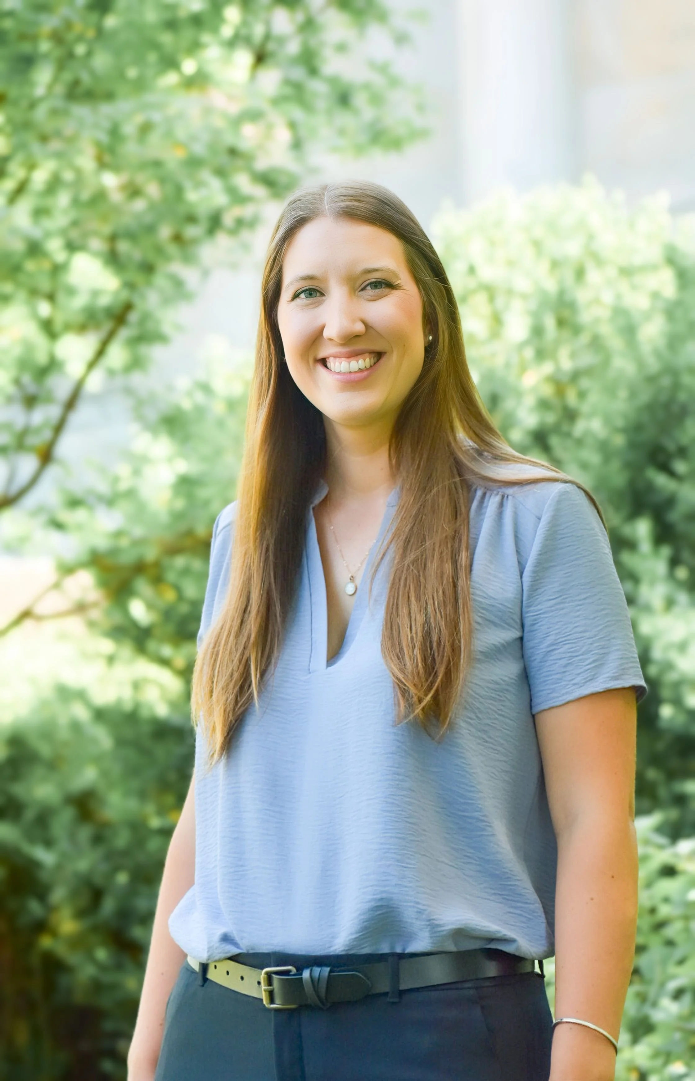 Portrait of a woman with long brown hair, wearing a light blue blouse and black pants, outdoors in a green, leafy setting, smiling at the camera.