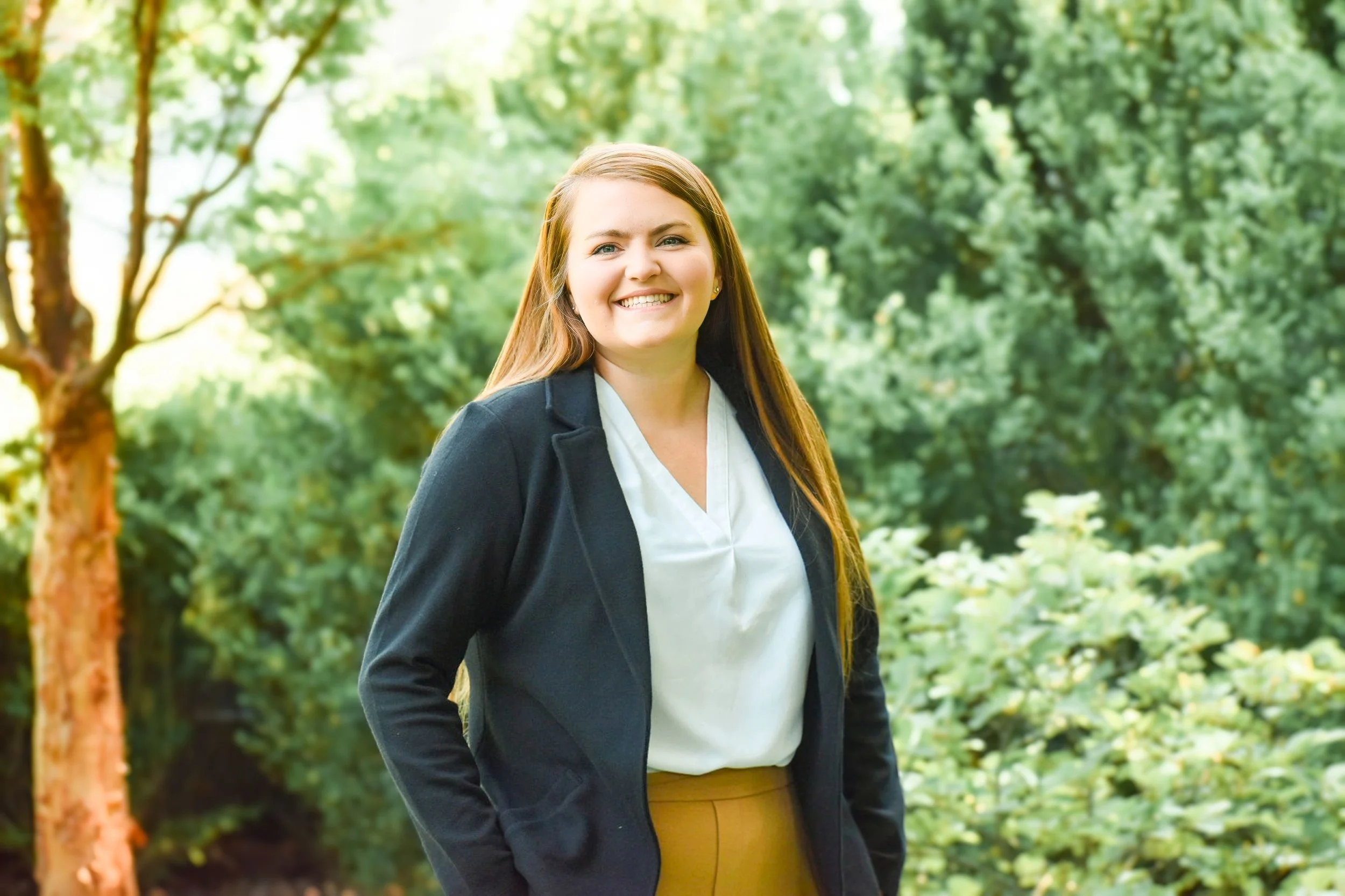 A woman smiling outdoors in a park with lush green trees and foliage in the background.