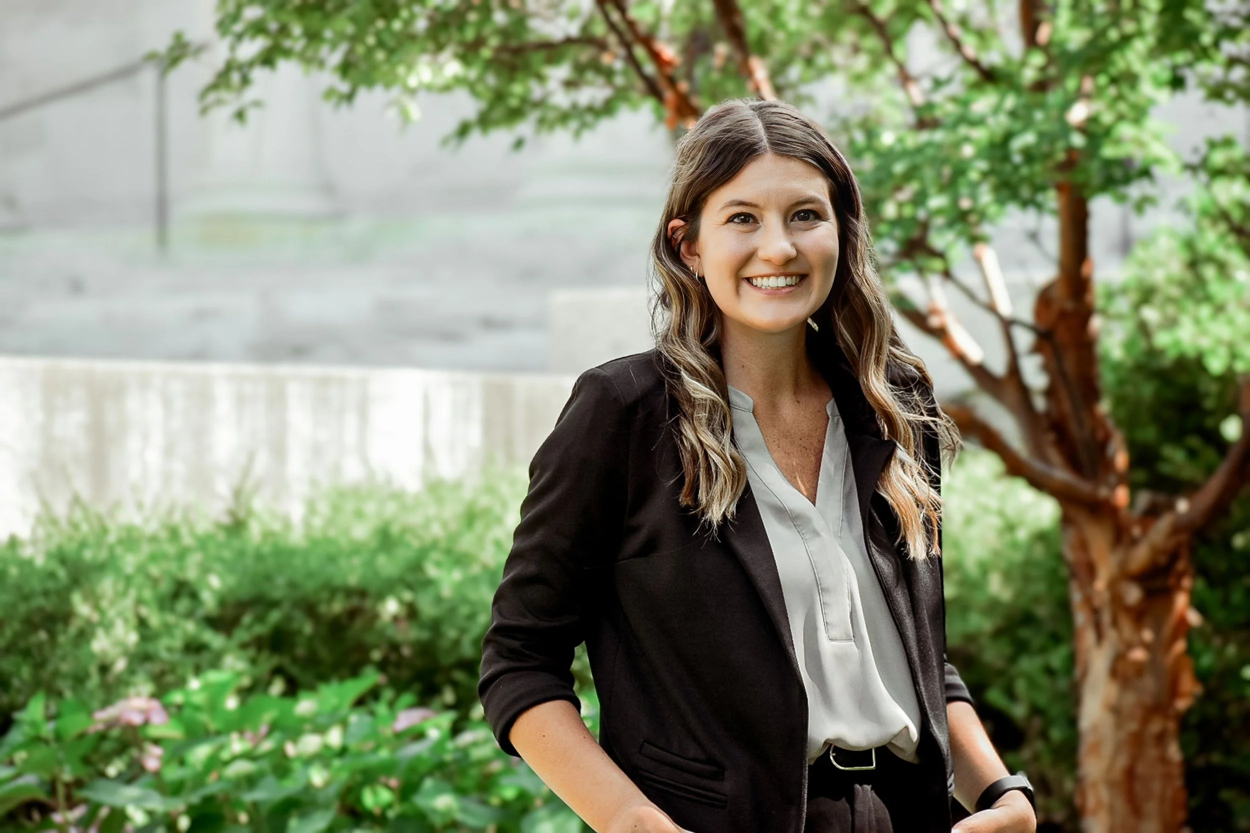 A smiling woman with long wavy brown hair standing outdoors in a garden with green plants and a tree in the background, dressed in a black blazer and light-colored blouse.