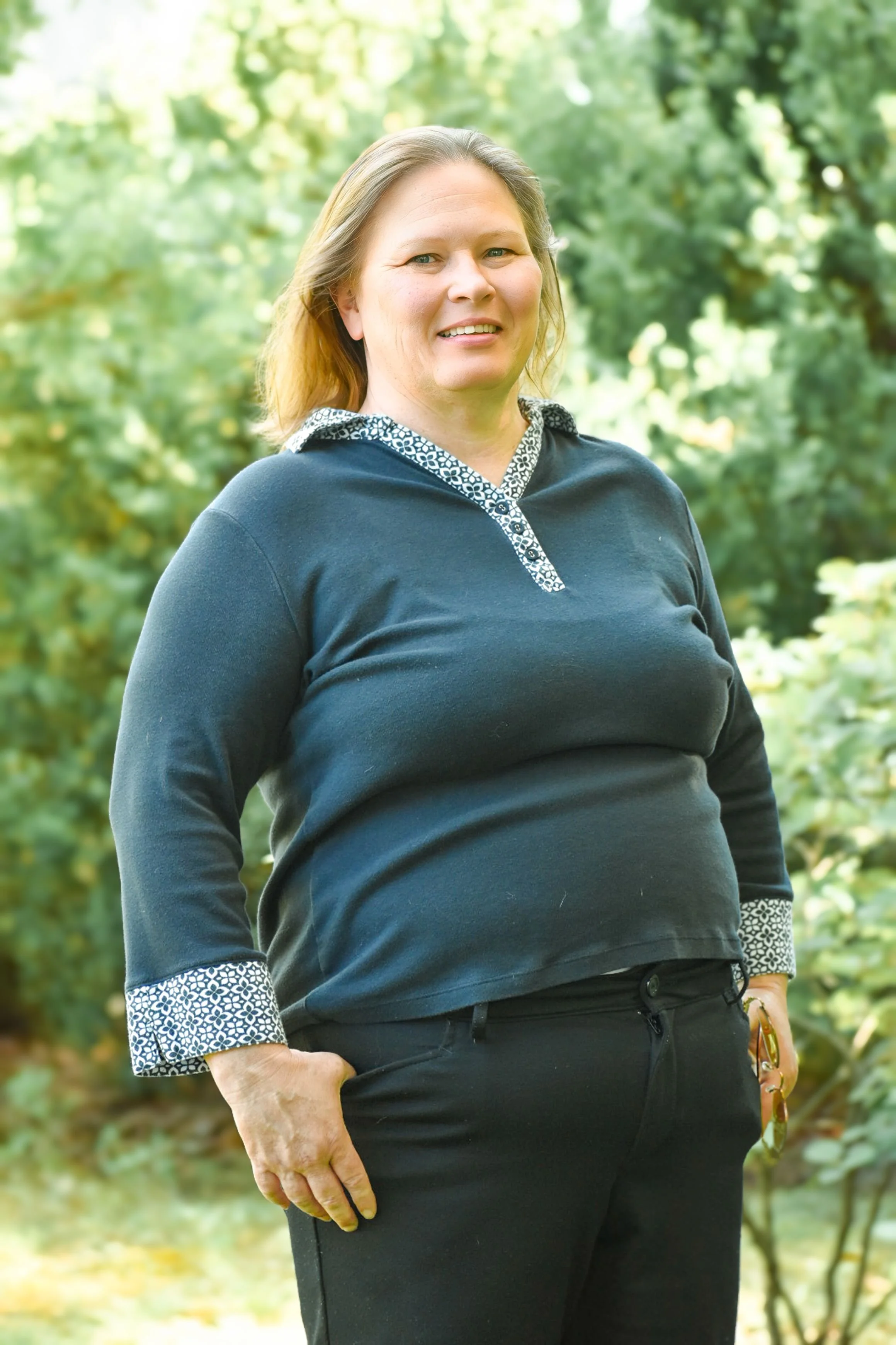 A woman standing outdoors in a natural setting with trees in the background, smiling at the camera. She is wearing a navy blue sweater with patterned cuffs and collar, and black pants.