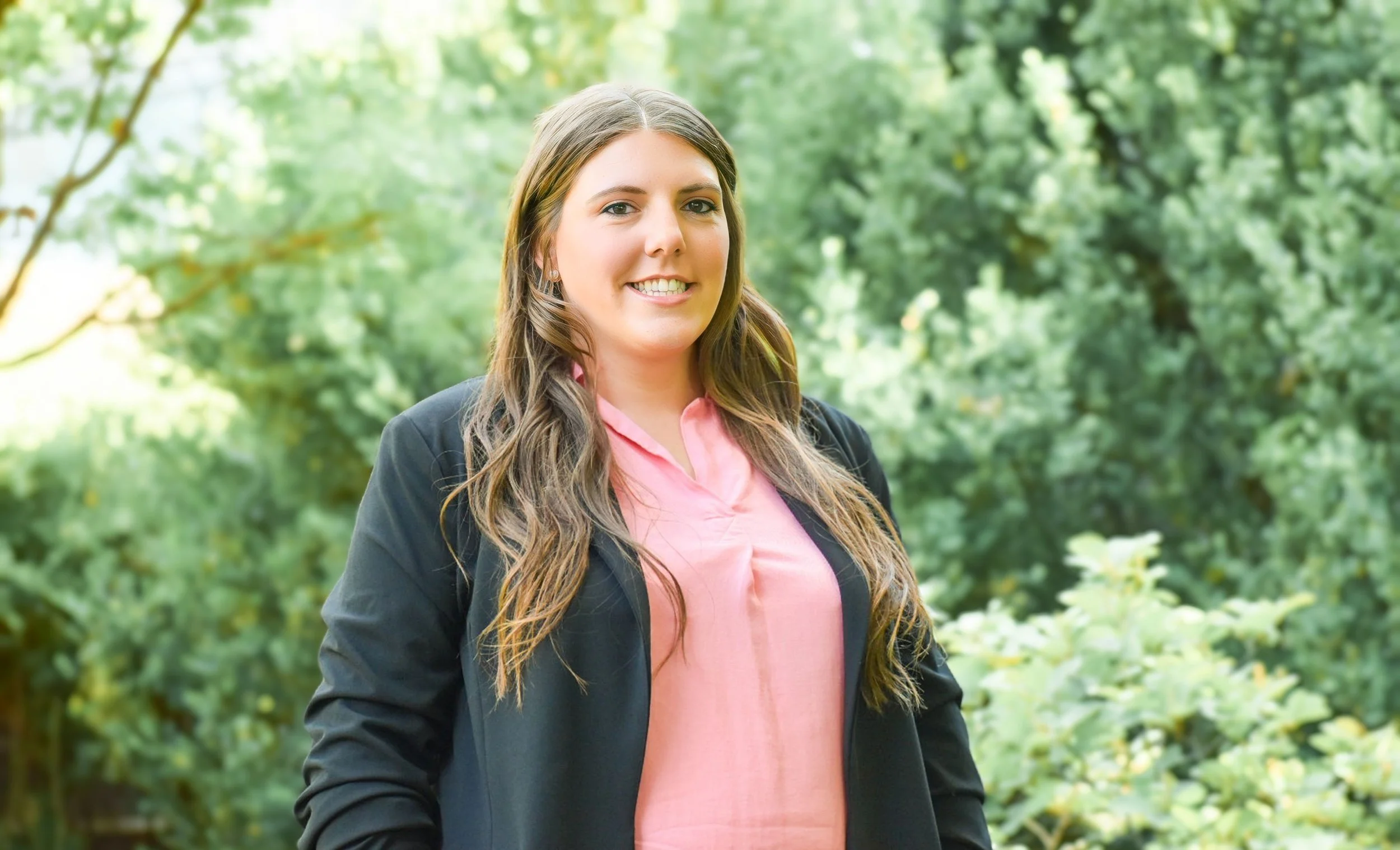 A woman with long brown hair smiling outdoors in a green park, wearing a pink blouse and black blazer.