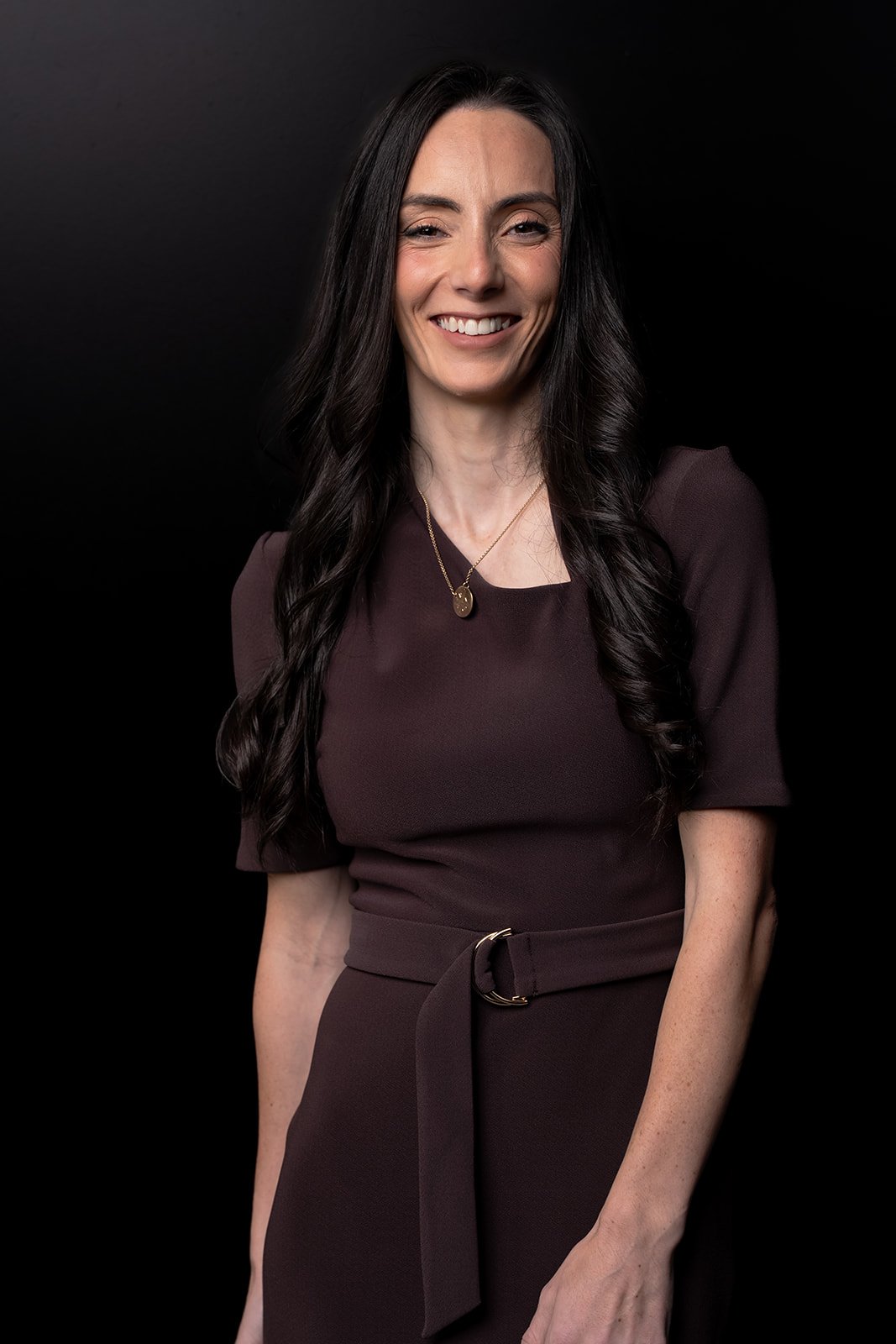 Smiling woman with long dark hair wearing a brown dress, against a black background.