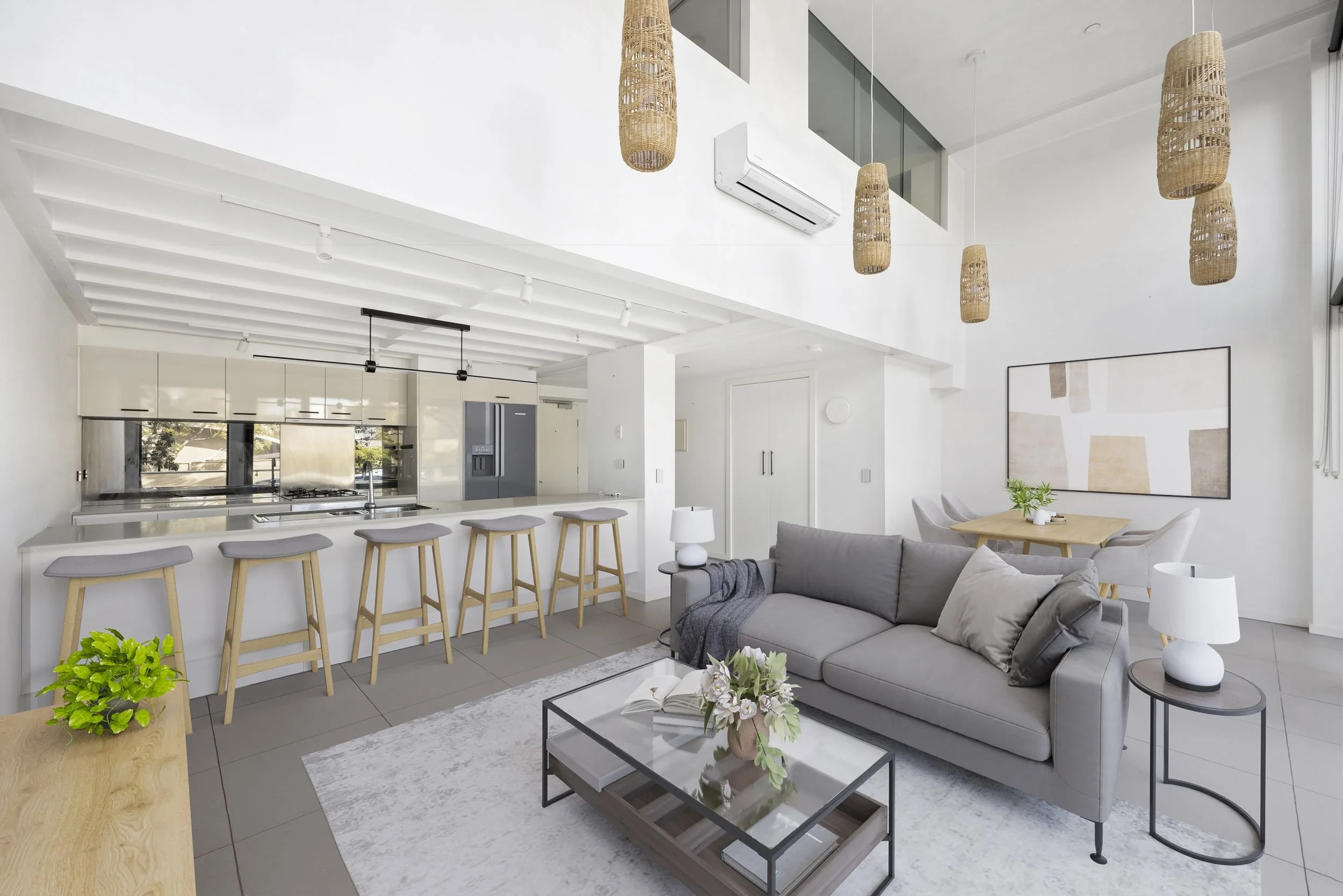 Modern, open-plan living room and kitchen with white walls and ceiling, pendant wicker lights, gray sofa, glass coffee table with flowers, dining table with chairs, kitchen with bar stools, and large window with natural light.