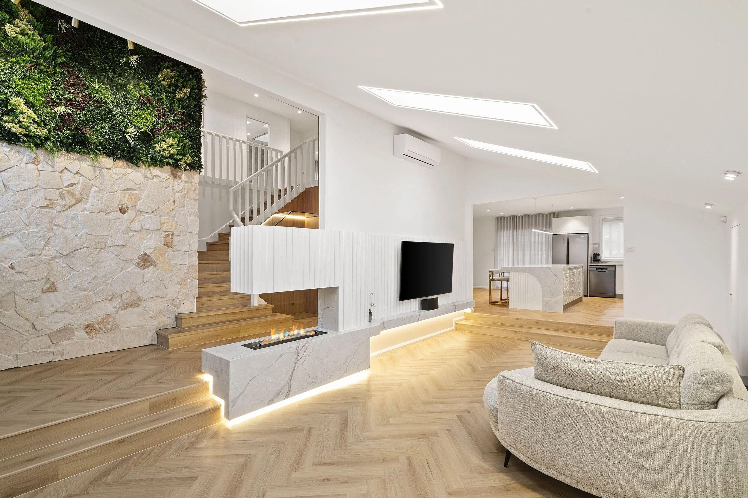 Modern living room with light wood floors, white walls, a built-in fireplace, a large curved sofa, and a flat-screen TV. The space features a staircase with white railing and a green plant wall. The kitchen area is visible in the background.