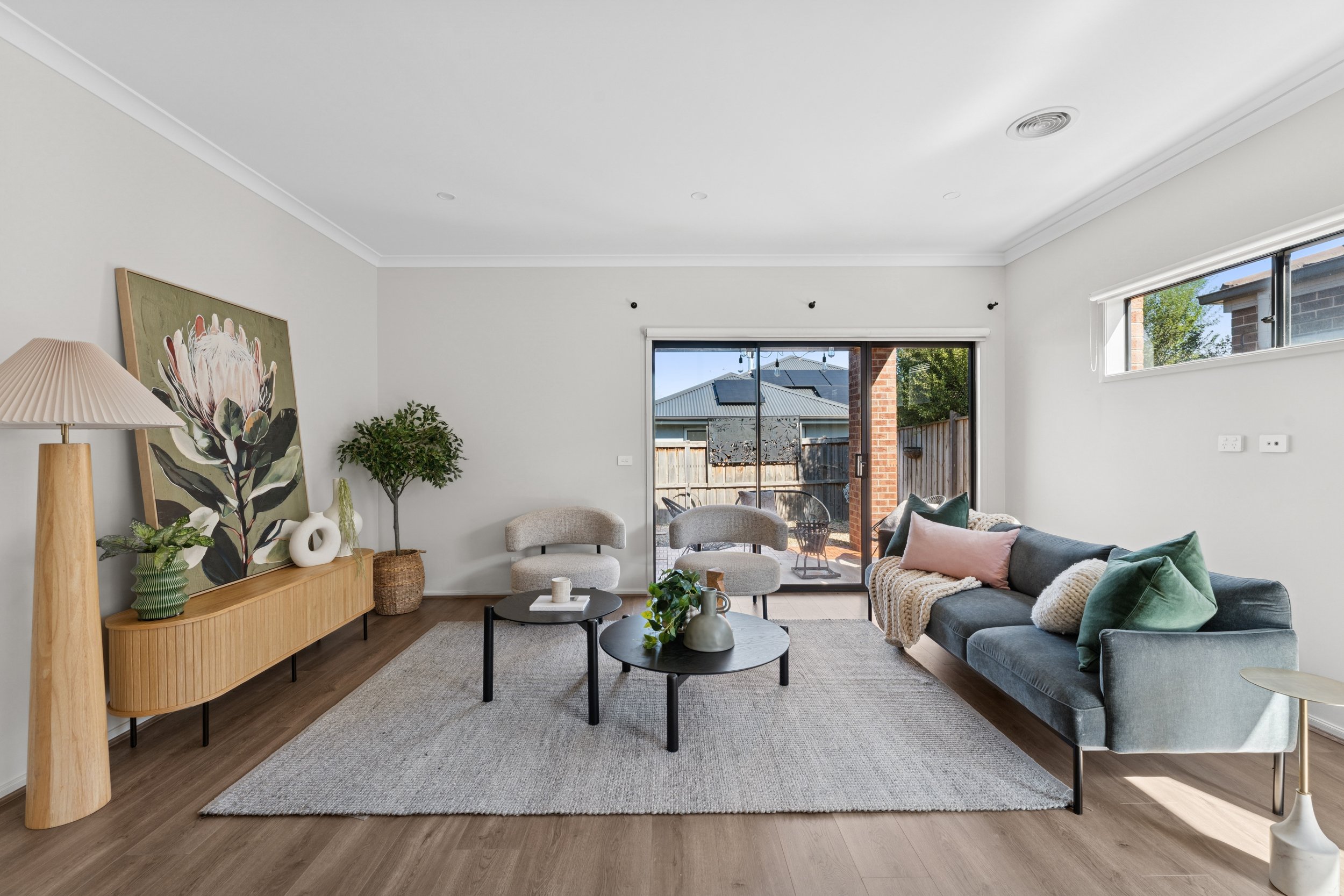 Modern living room with a gray sofa, pastel pillows, a textured rug, black coffee tables, and a sliding glass door leading to an outdoor patio.