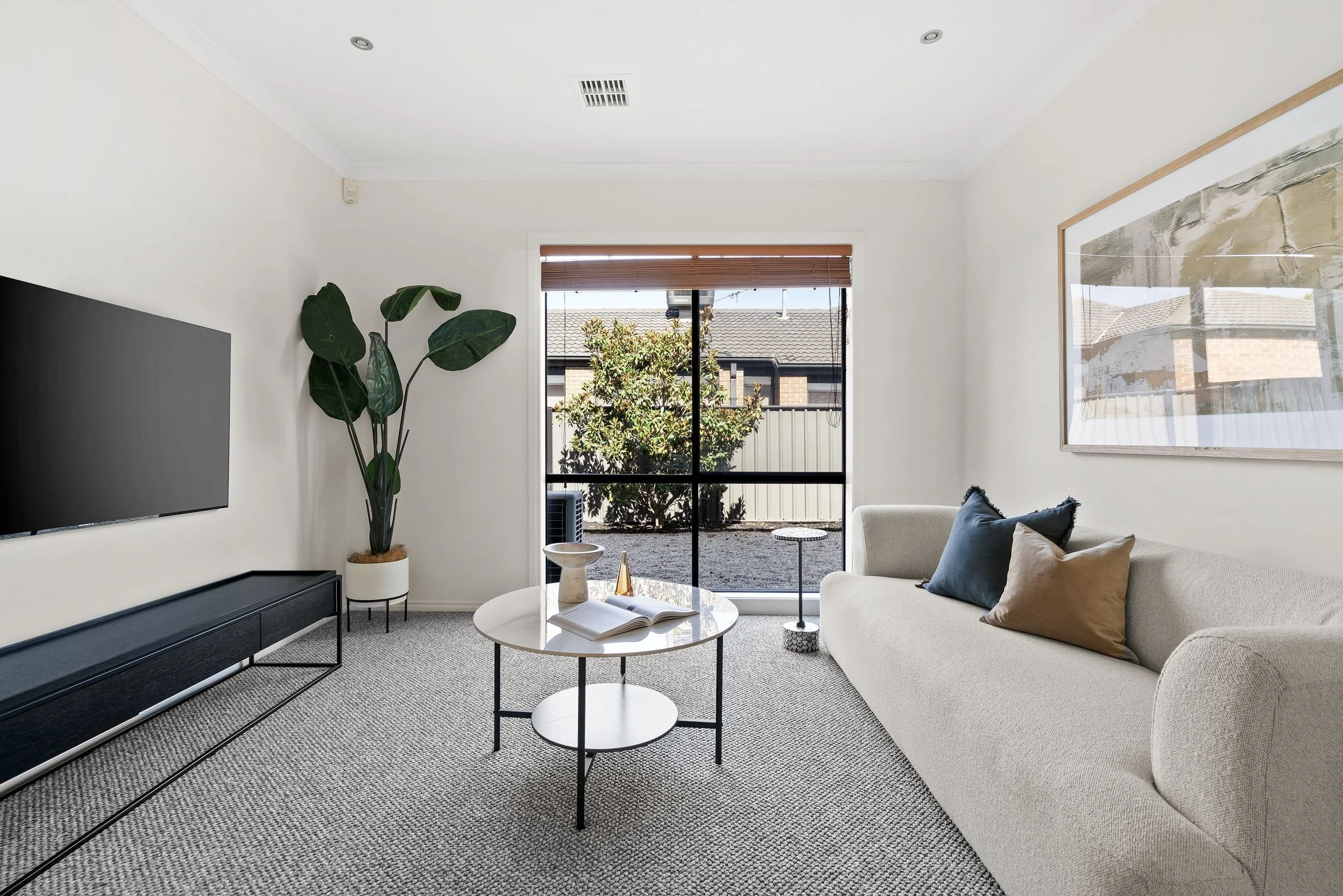 A minimalist living room with a beige sofa, a black TV mounted on the wall, a tall potted plant, a round coffee table with books and decorations, a large window with a view of a backyard, and framed artwork on the wall.