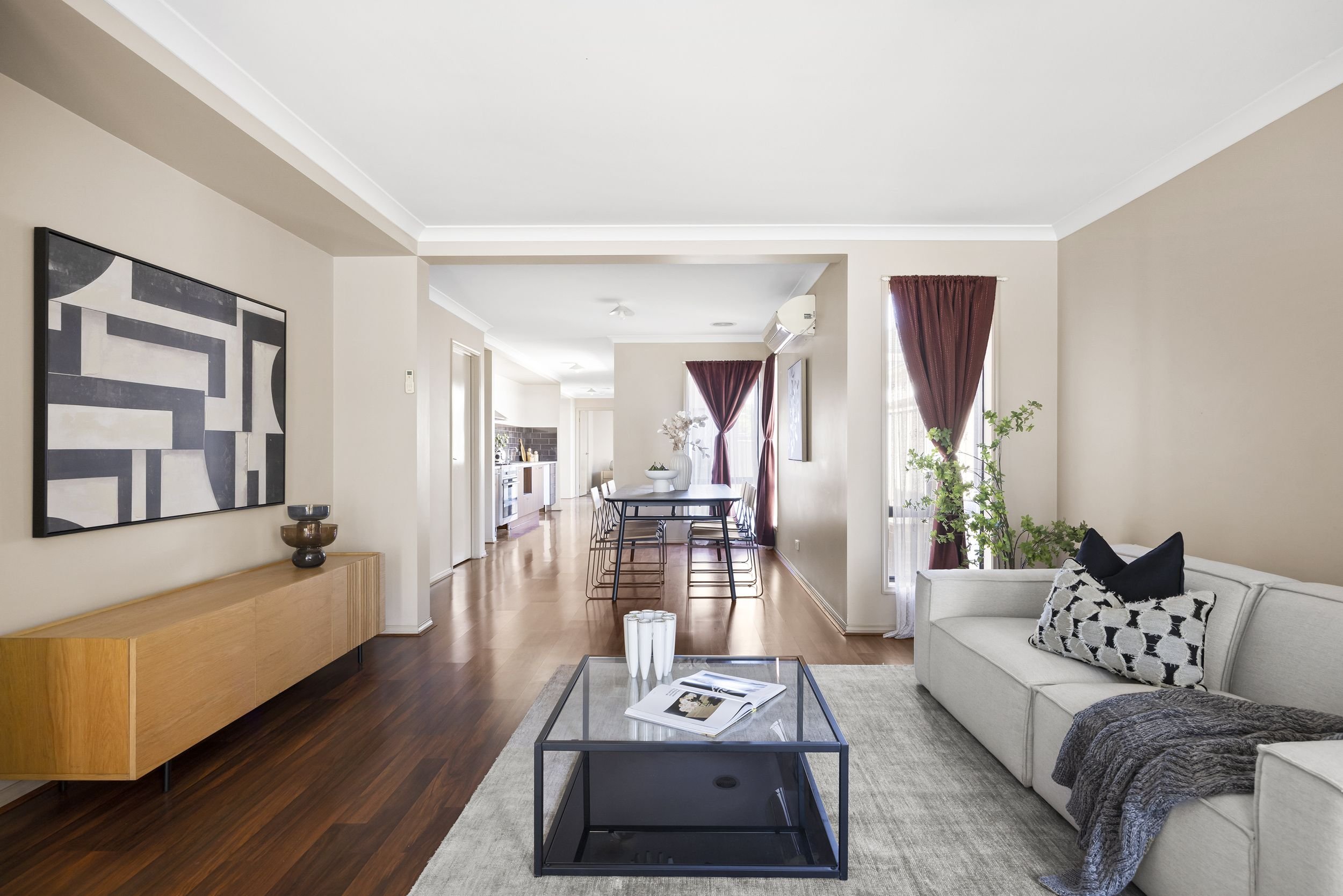 Living room with a white couch, black and white pillows, a glass coffee table, and a beige rug. Artwork on the wall, wooden sideboard, and a few potted plants. Door to a dining area with a table and chairs, red curtains, and a kitchen in the background.