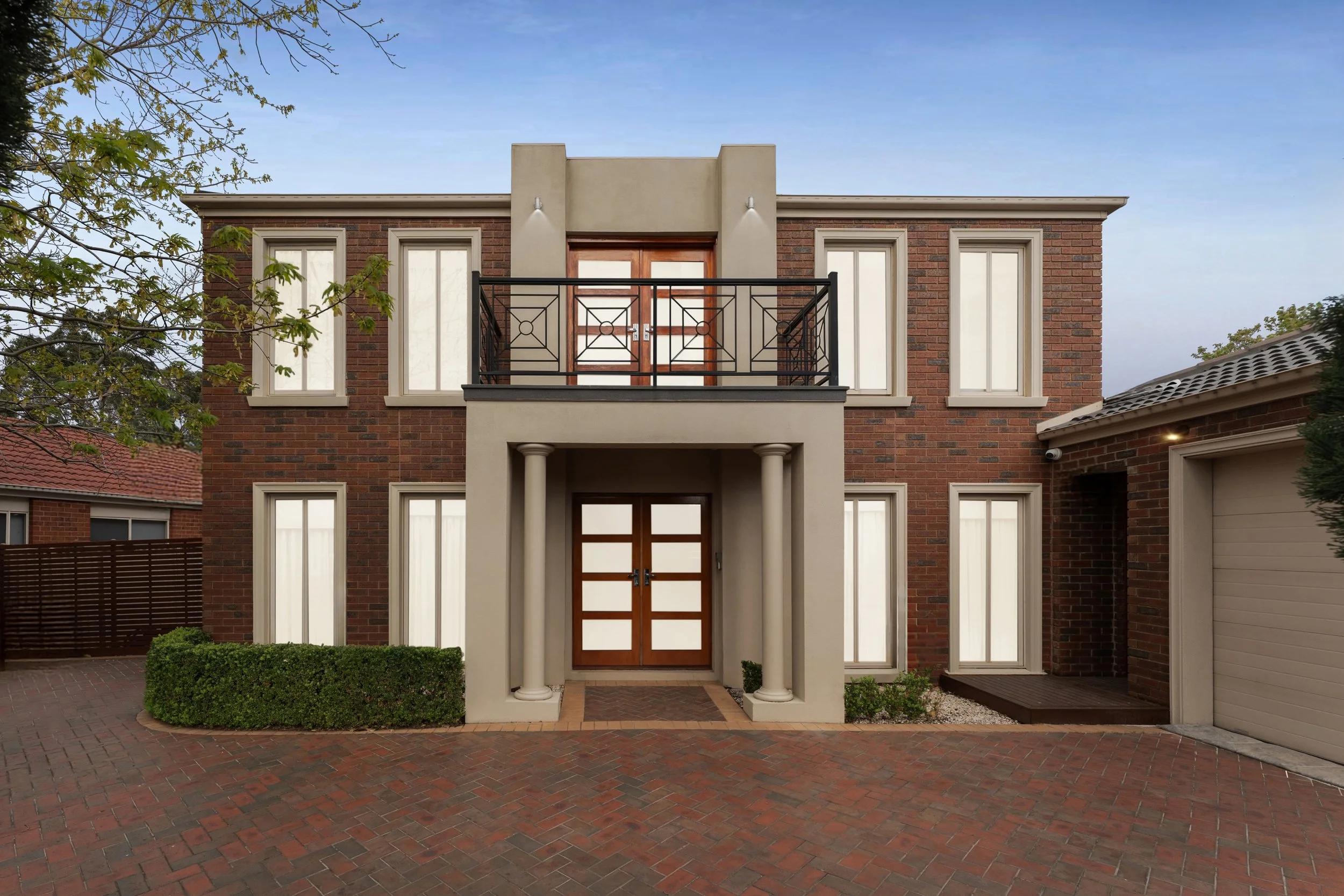 Front view of a two-story brick house with a symmetrical design, featuring a central entrance with a covered porch supported by two columns, and a balcony with a black railing above it. The house has multiple tall, narrow windows with cream-colored frames and white curtains. The driveway is paved with red bricks, and there is a small neatly trimmed hedge in front of the porch.