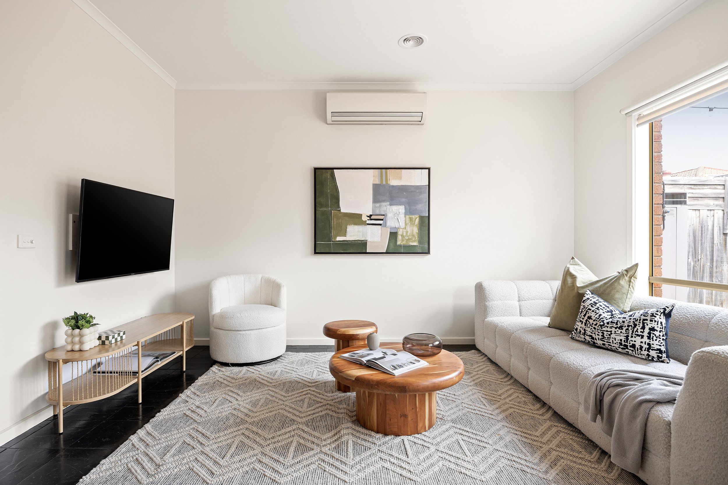 Living room with white walls, black hardwood floors, a flat-screen TV on the left wall, a light-colored armchair, a wooden coffee table with magazines, a white sofa with decorative pillows, a large window, and minimal wall art.
