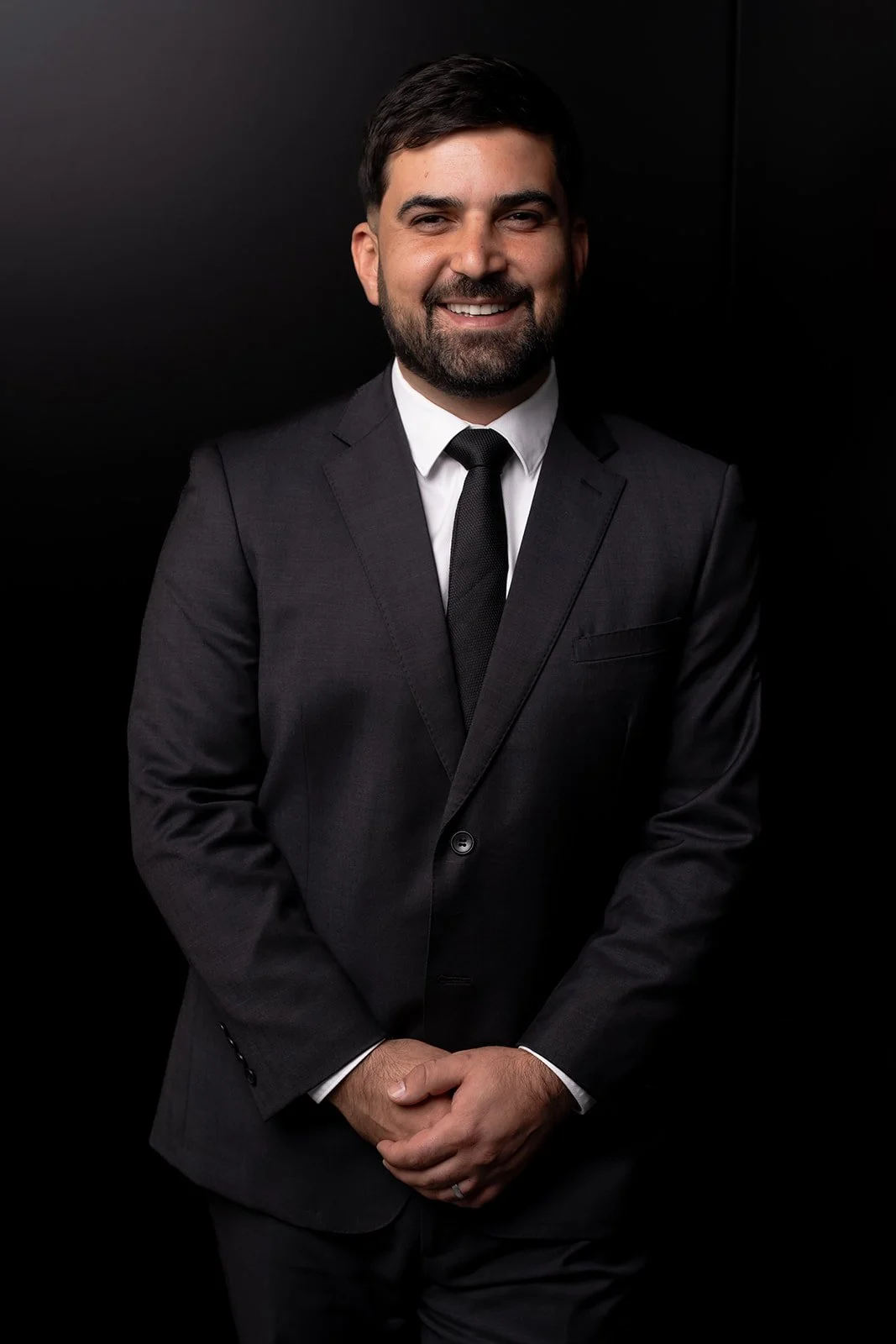 Smiling man in black suit and tie with black background.